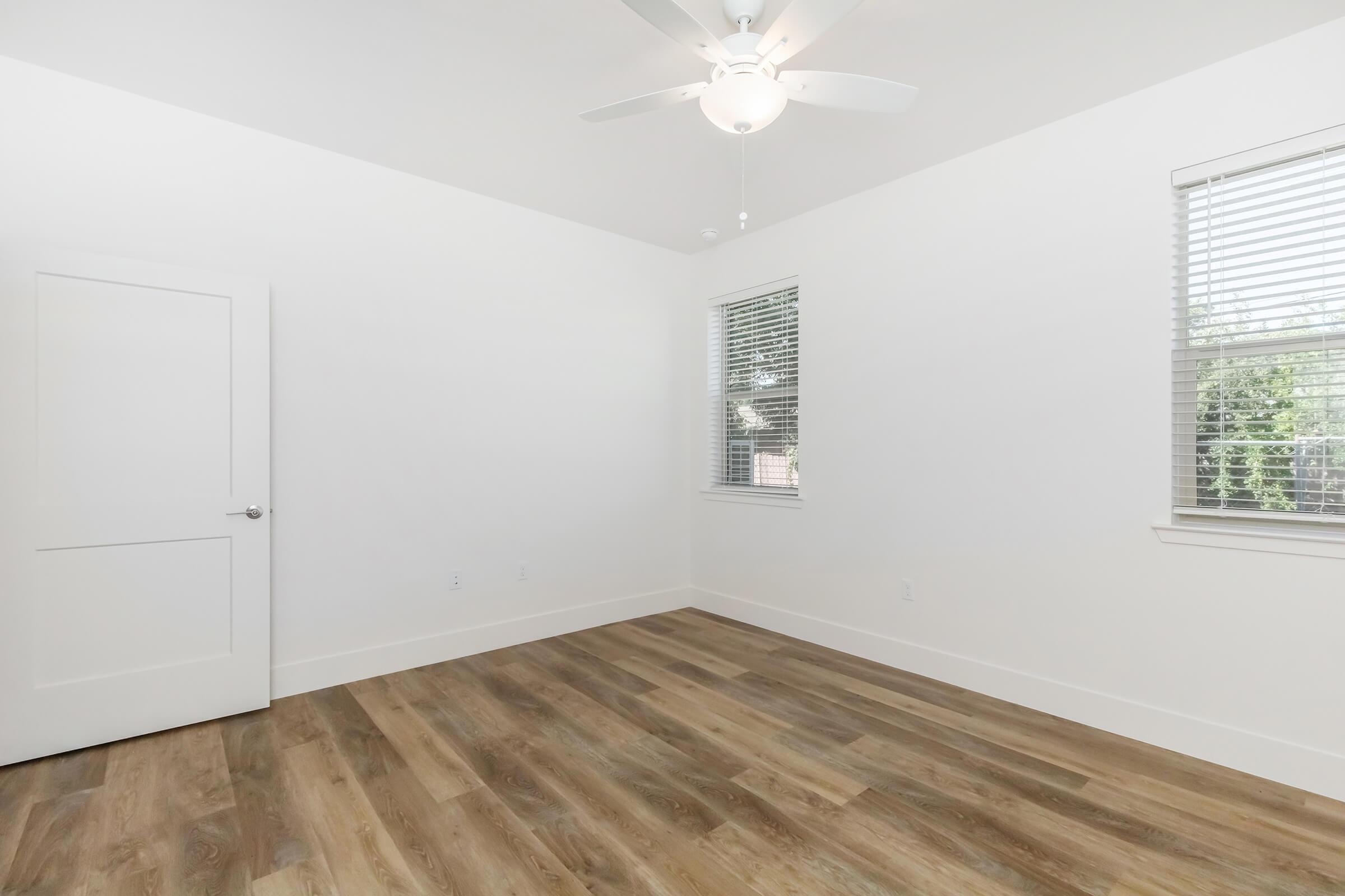 A bright and spacious room featuring light wood flooring, a ceiling fan, and two windows with blinds. The walls are painted white, creating a clean and airy atmosphere. A white door leads to another area, enhancing the modern look of the space. Ideal for a bedroom or office.