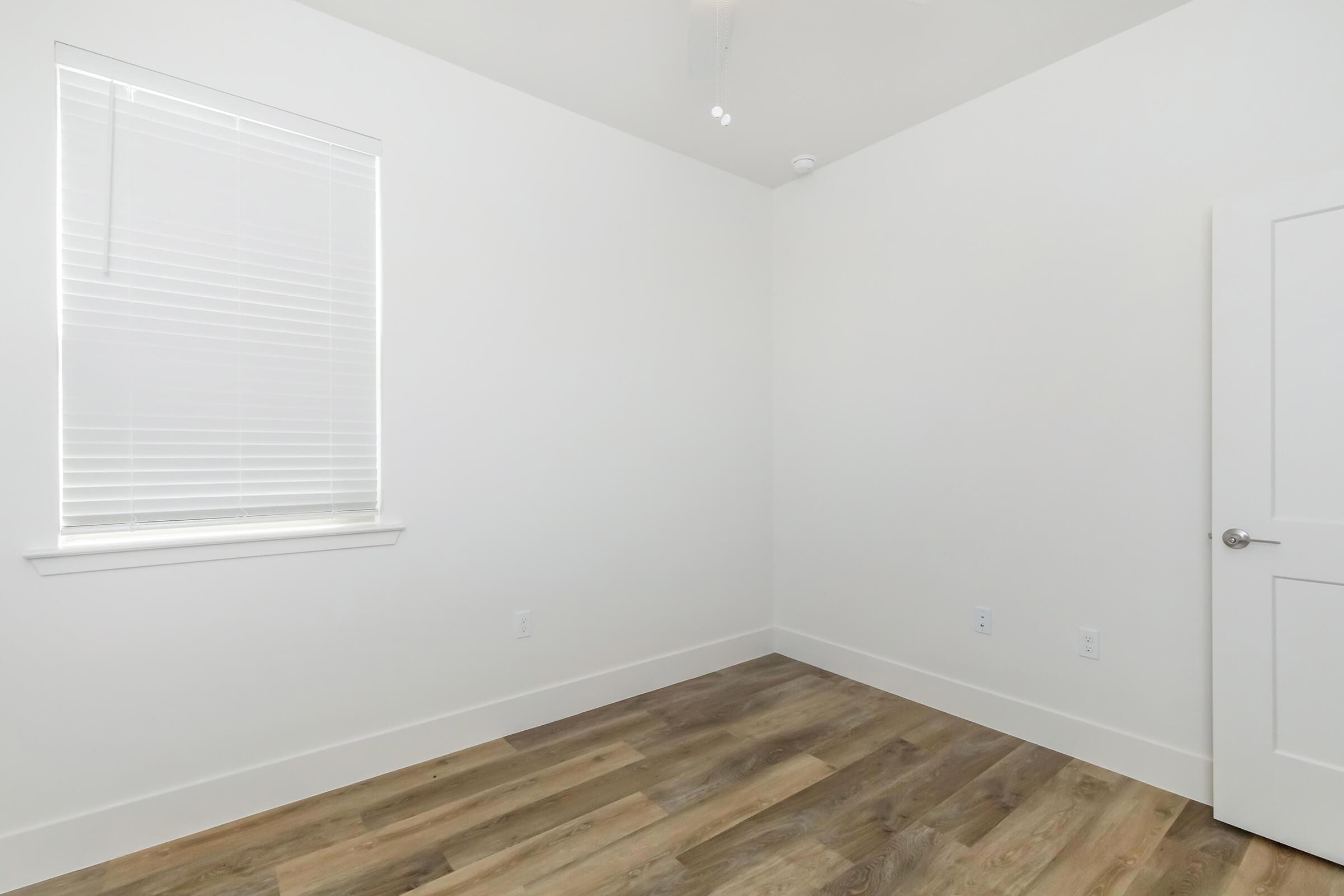 A vacant room with light-colored walls and a single window featuring white blinds. The floor is made of wooden planks, and there is a plain white door on one side. The space is empty, creating a clean and minimalistic look.