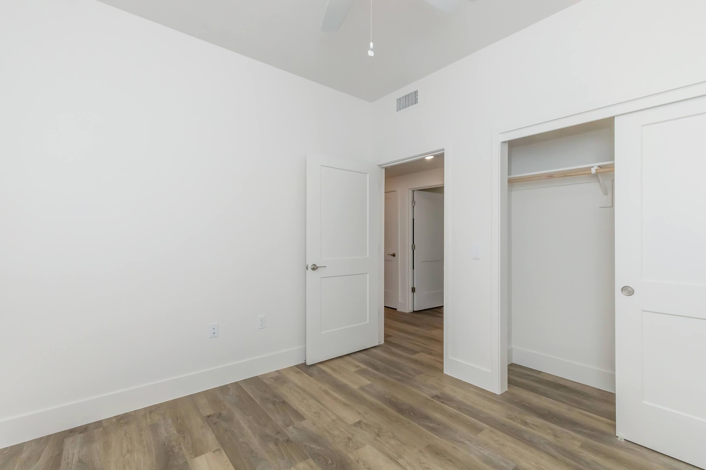 A spacious interior view of a light-colored room featuring a white ceiling fan, beige flooring, and two doors: one leading to a closet and the other to a hallway. The walls are undecorated, creating a bright and airy atmosphere.