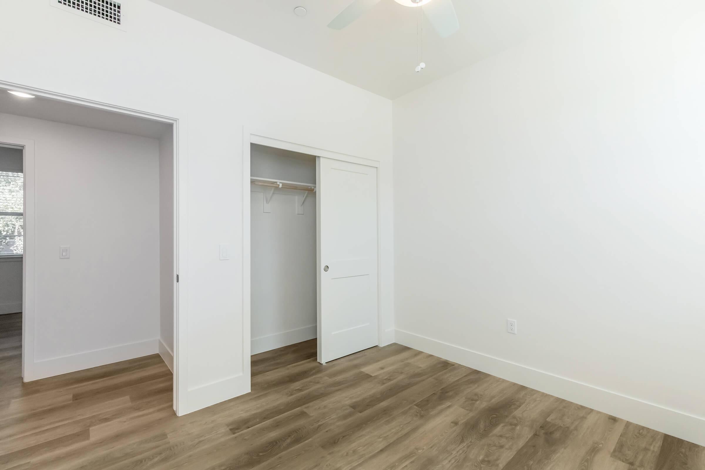 A bright, minimally furnished room with white walls and light wooden flooring. There is an open closet with a sliding door, a ceiling fan, and a doorway leading to another space. Natural light enters through a nearby window, creating an airy atmosphere.