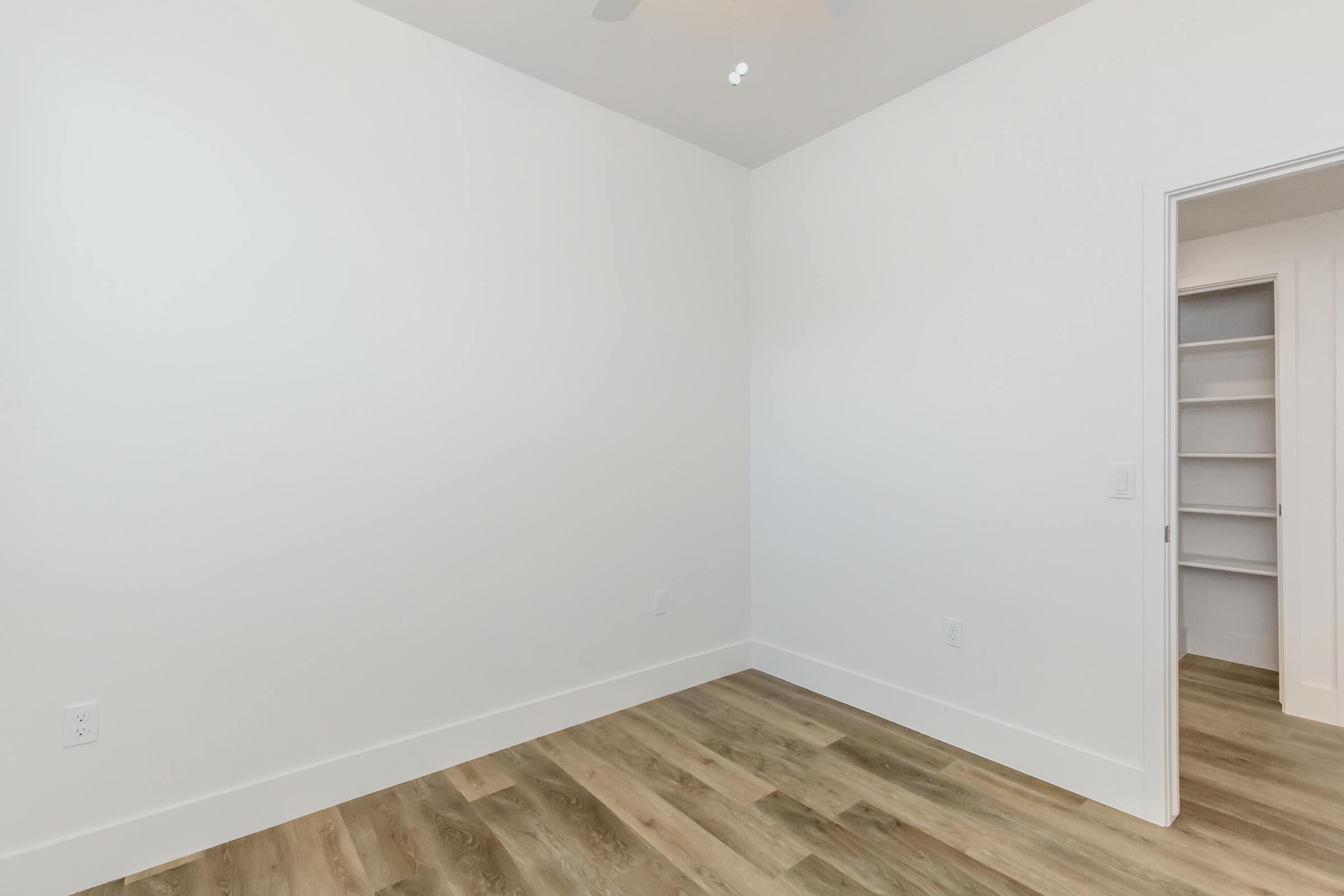 Empty room with white walls and light wood flooring, featuring a ceiling fan. A doorway leads to a closet with shelves, and natural light enters from an unseen window, creating a bright, minimalist atmosphere.