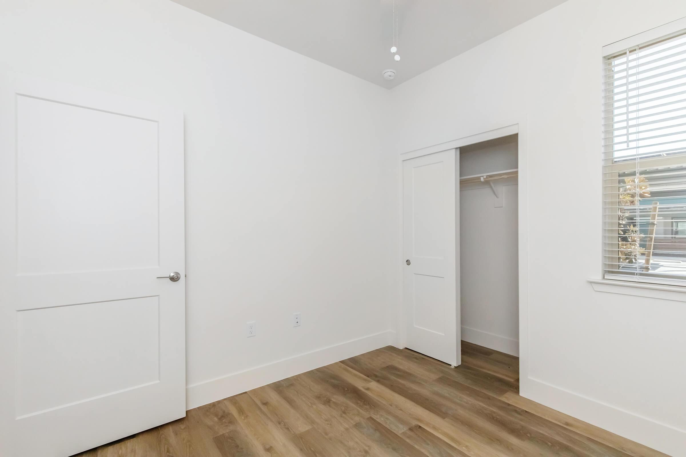 Empty room featuring white walls, a light-colored wooden floor, a closet with sliding doors, and a ceiling fan. Two doors can be seen on the left side, leading outside the room and into the closet. Natural light streams in through a window with blinds.