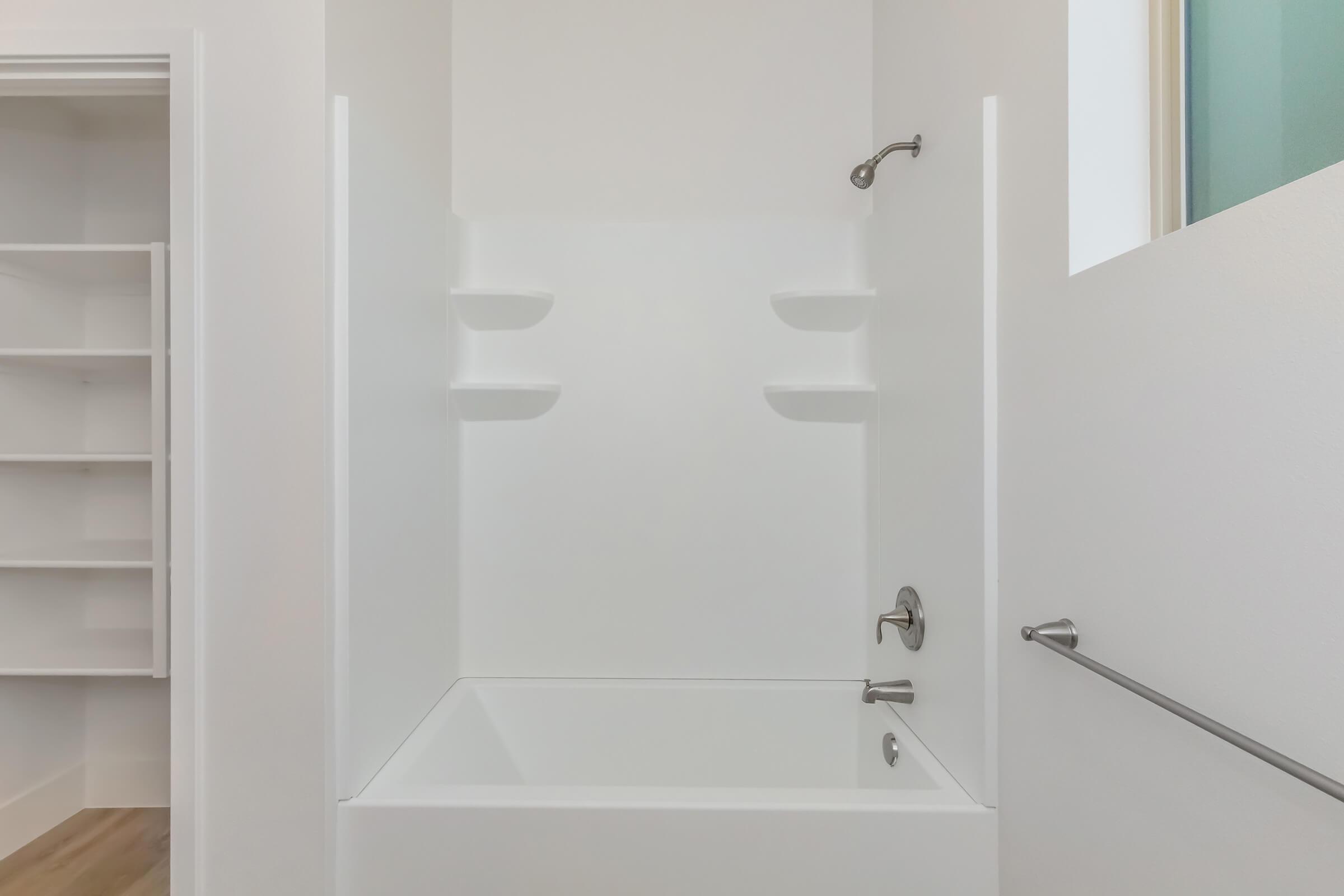 A clean, modern bathroom featuring a white bathtub with built-in shelves on the wall. The bathtub is designed for a shower and is situated in a bright, minimalist space with light-colored walls and wooden flooring. A door partially open reveals a glimpse of a storage area.