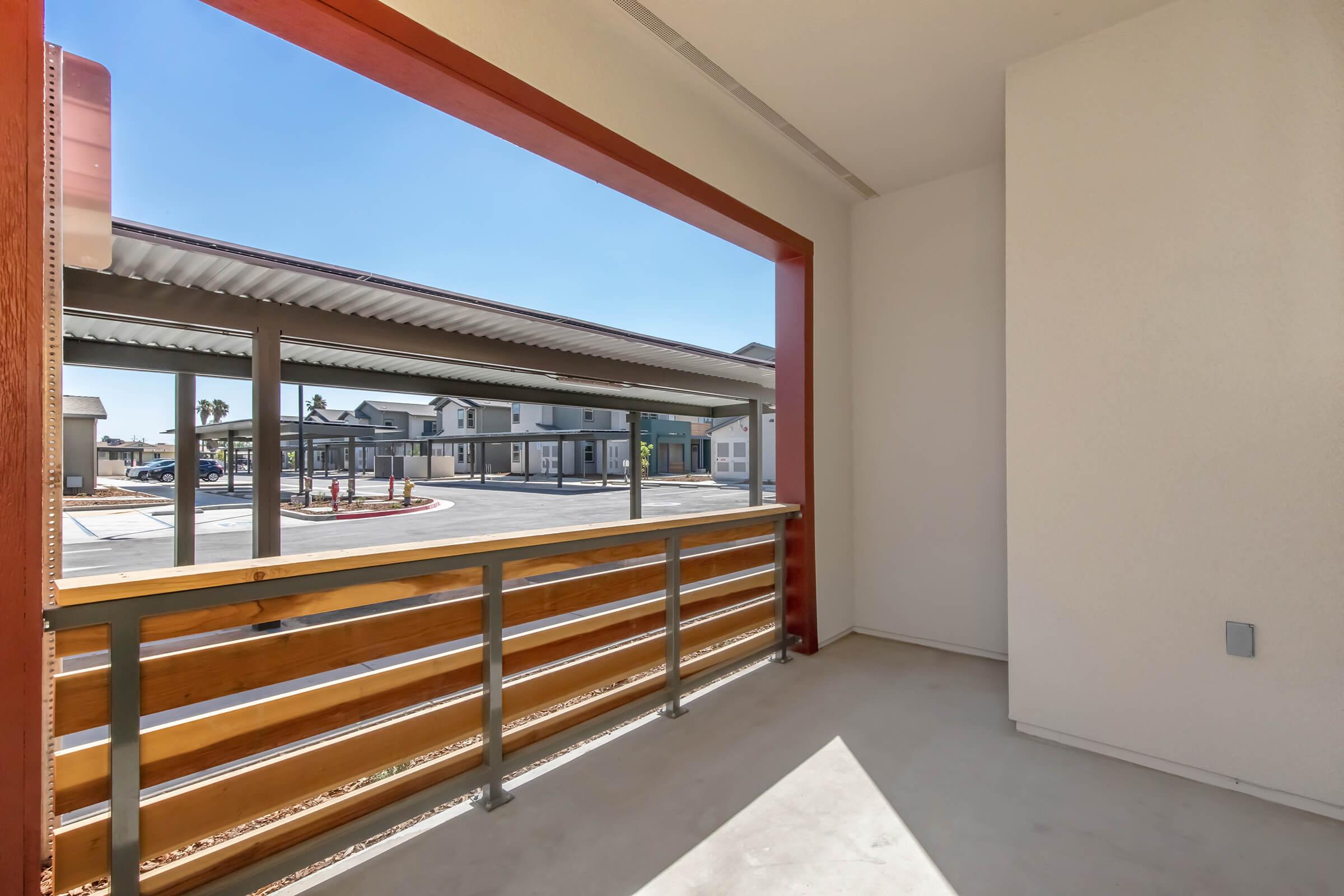 A view from a balcony featuring a wooden railing. The scene shows a clear blue sky and a neighboring building with a parking area visible below. The balcony space is bright and spacious, with clean lines and minimal decor.