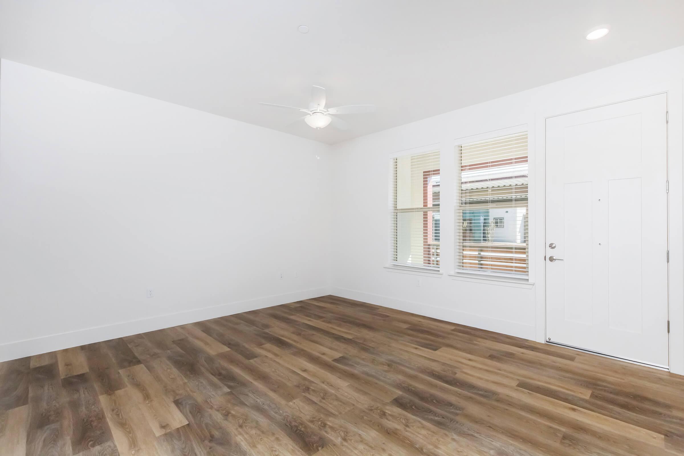 A bright, empty room with white walls and a ceiling fan. The floor features light brown wooden planks. There are large windows allowing natural light, and a front door with a simple design is visible. The overall atmosphere is clean and modern, ideal for various interior design possibilities.