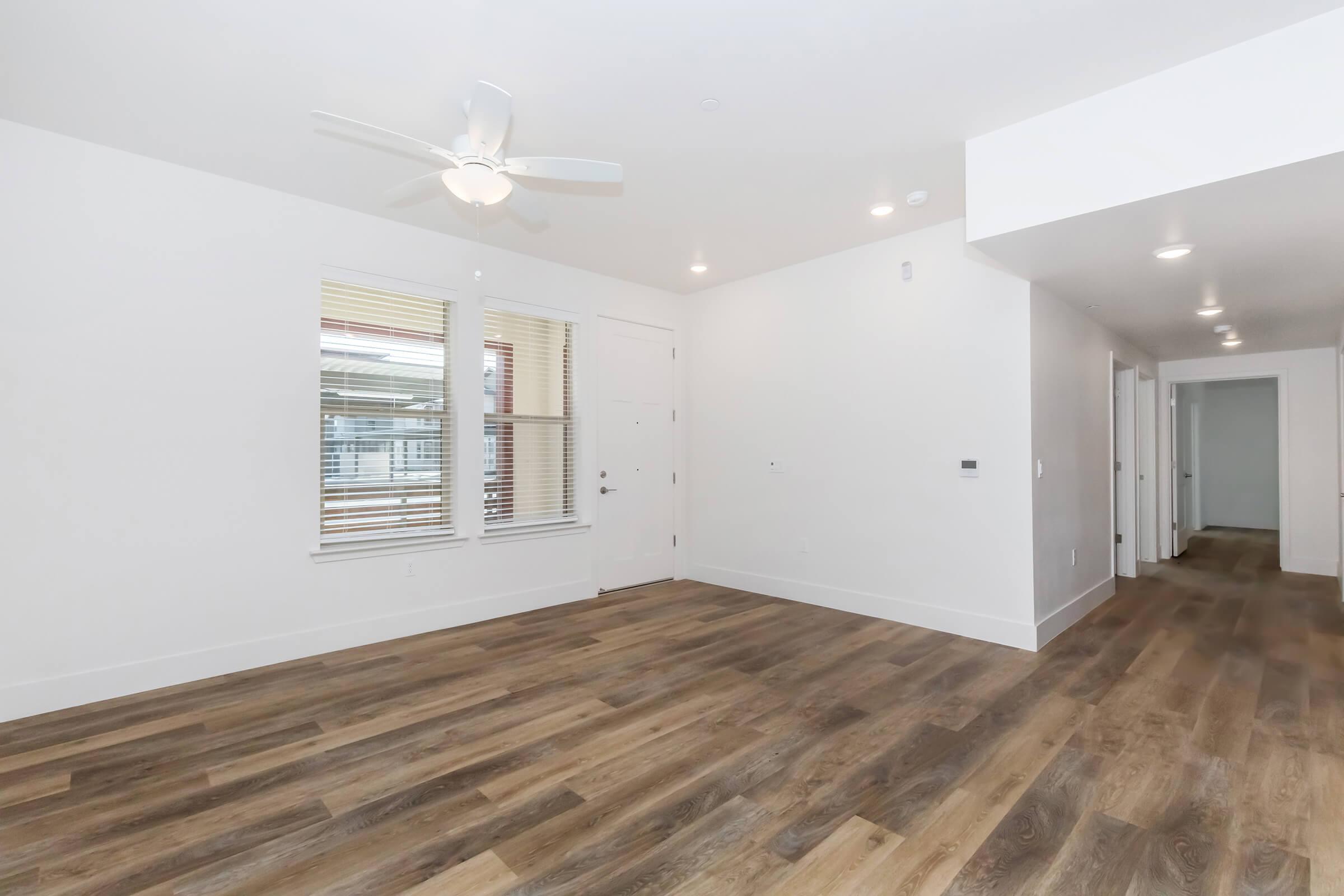 A spacious, modern living area featuring light-colored walls, a ceiling fan, and large windows with blinds. The flooring is a warm-toned hardwood, and the space has an open layout that leads to a hallway with doors, providing a bright and inviting atmosphere.