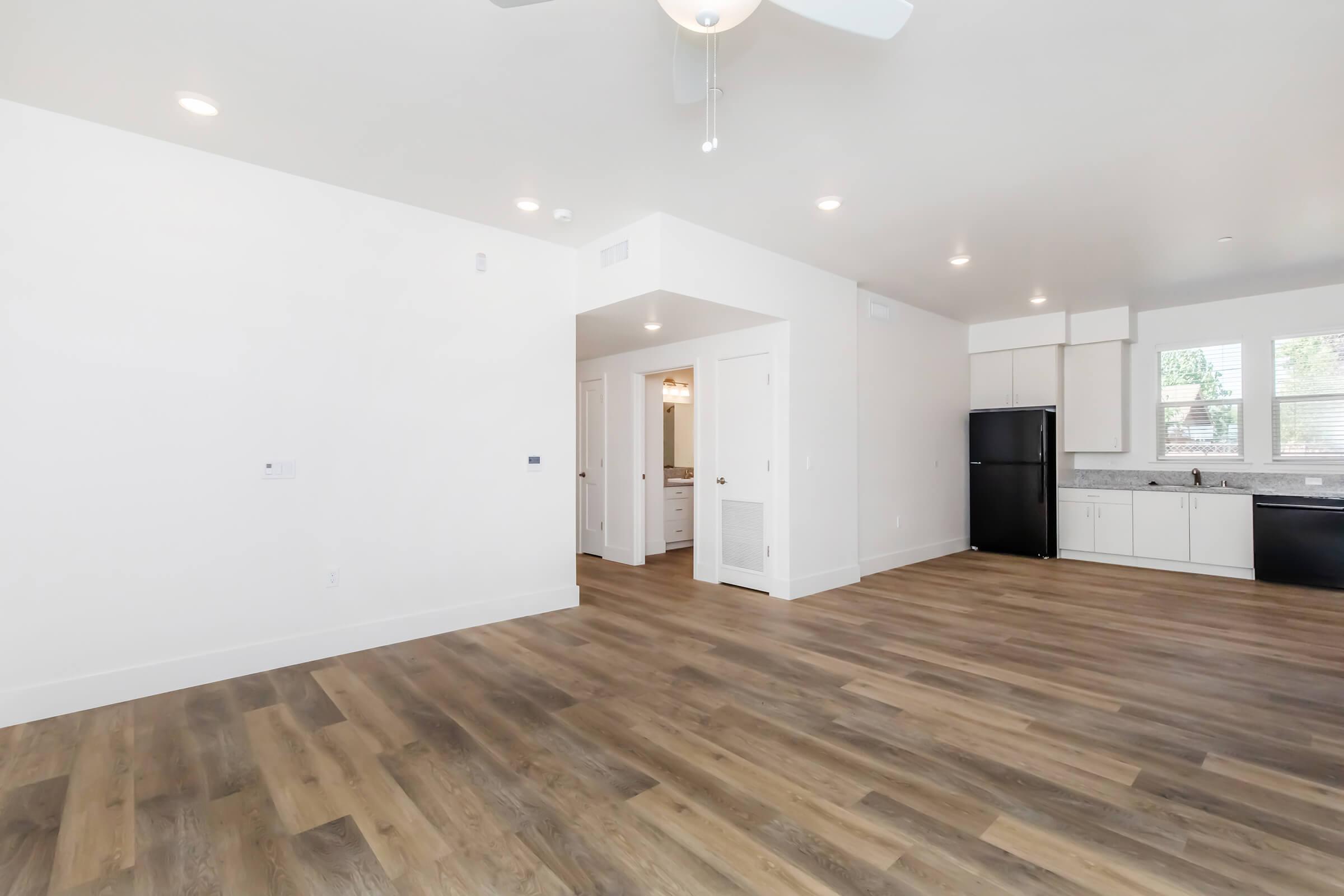 Spacious and modern living area with light-colored walls and wooden flooring. Features include a ceiling fan, an open layout, and a kitchen area with white cabinetry and a dark refrigerator. Bright natural light enters through windows, enhancing the contemporary feel of the space.