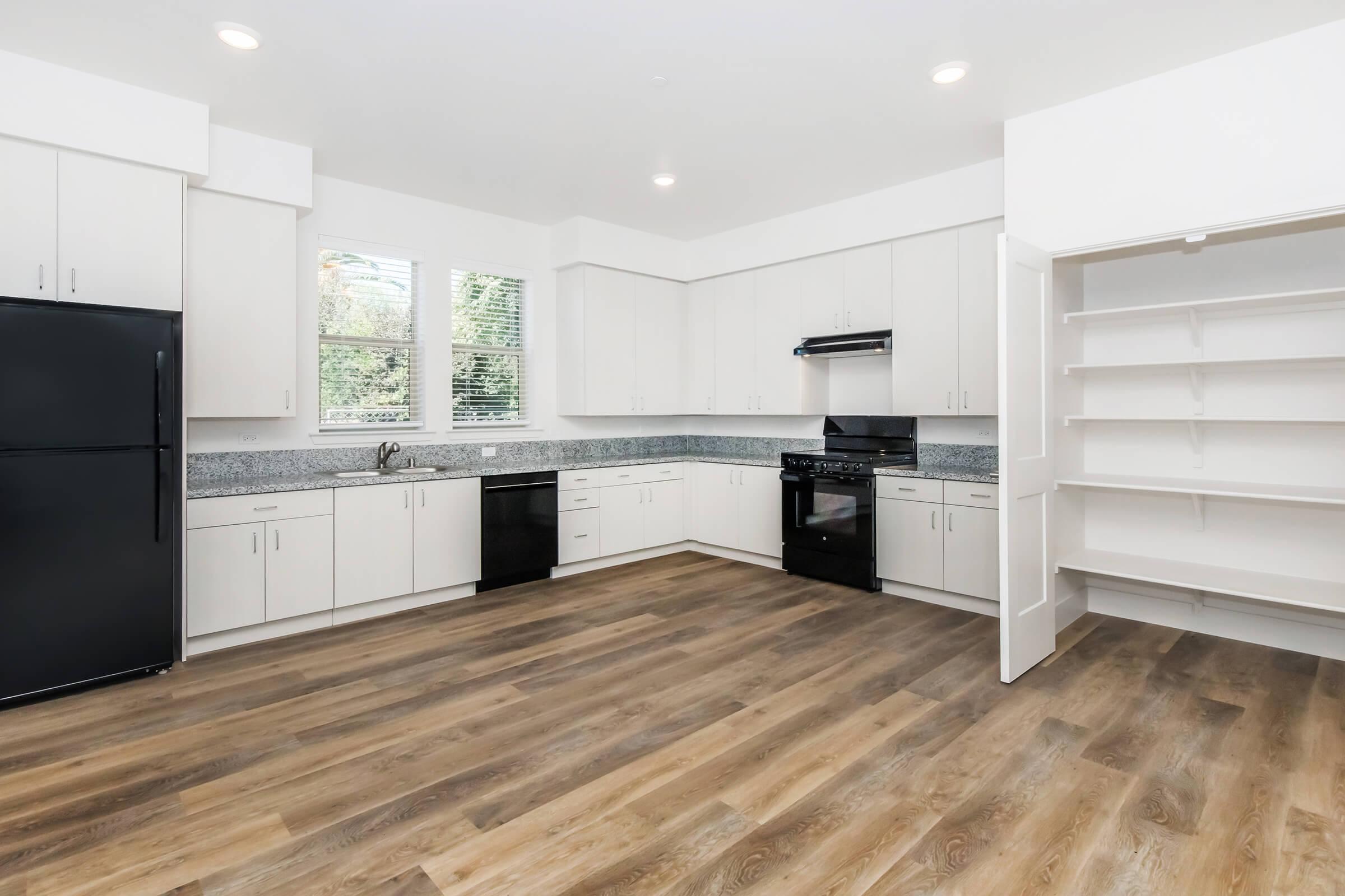 A modern kitchen with white cabinetry, a black refrigerator, an oven, and a stove. The countertops feature a gray speckled design, and large windows provide natural light. There is a spacious open layout with wooden flooring and an empty pantry cupboard on the right.
