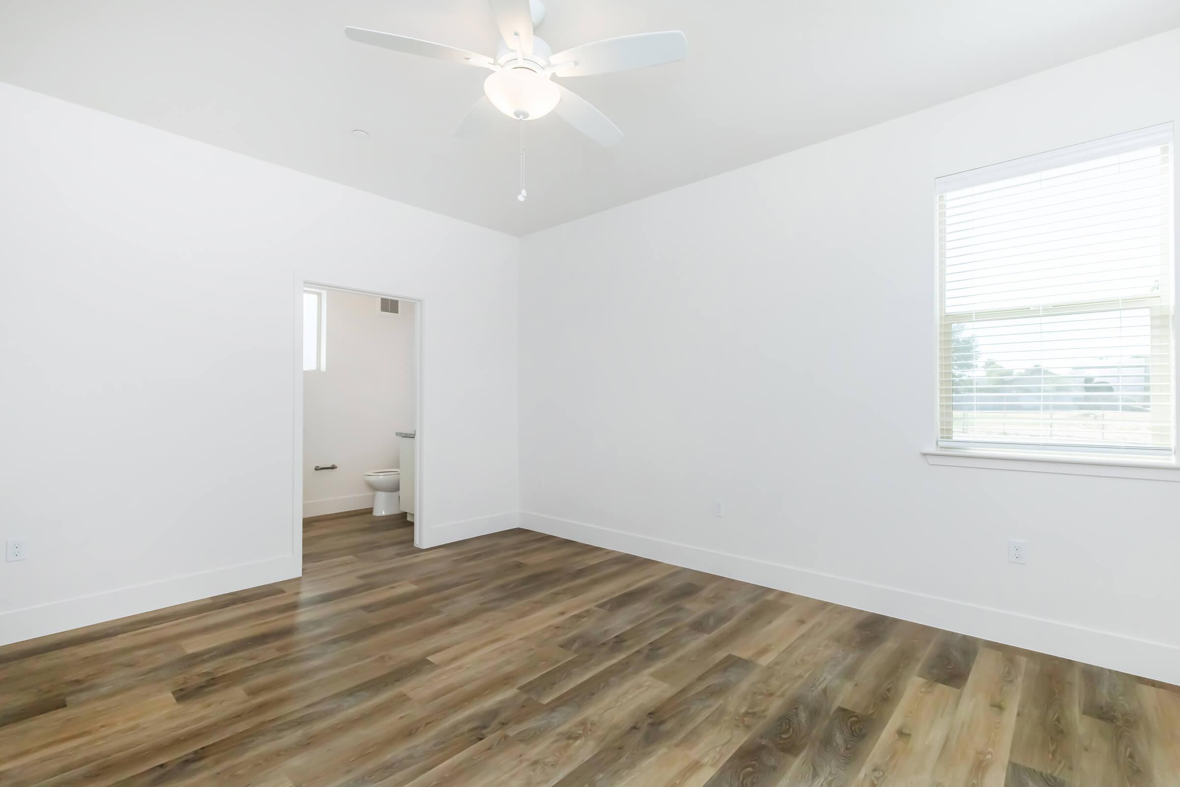 A bright, spacious room with white walls and hardwood flooring. A ceiling fan hangs from the ceiling, and there's a doorway leading to a small bathroom. A window on one wall lets in natural light, illuminating the clean and minimalist design of the space.