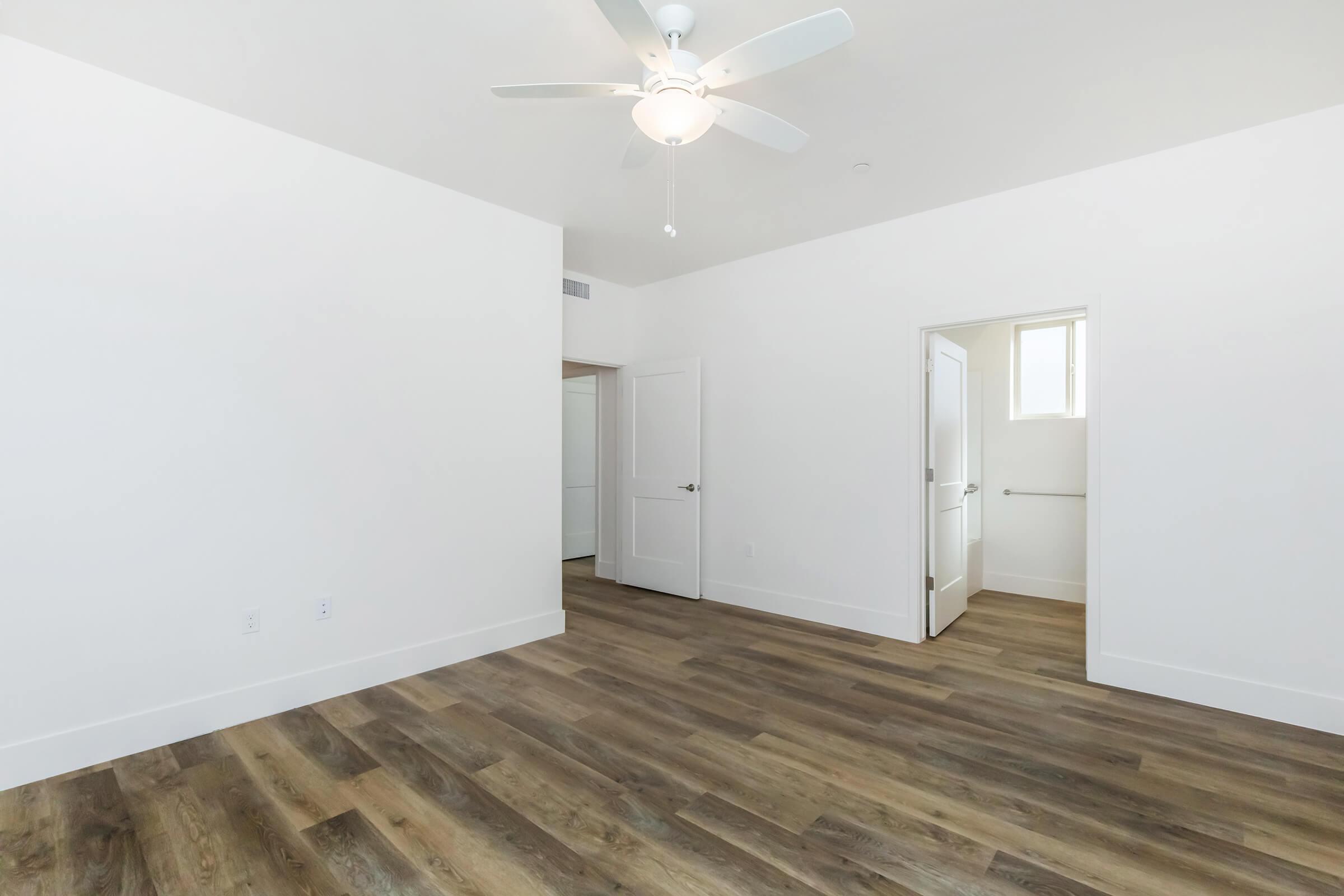 A bright, empty room with light wood flooring and white walls. It features a ceiling fan and an open door leading to another room. Natural light enters through a window, creating a spacious and airy feel. The overall design is modern and minimalist.