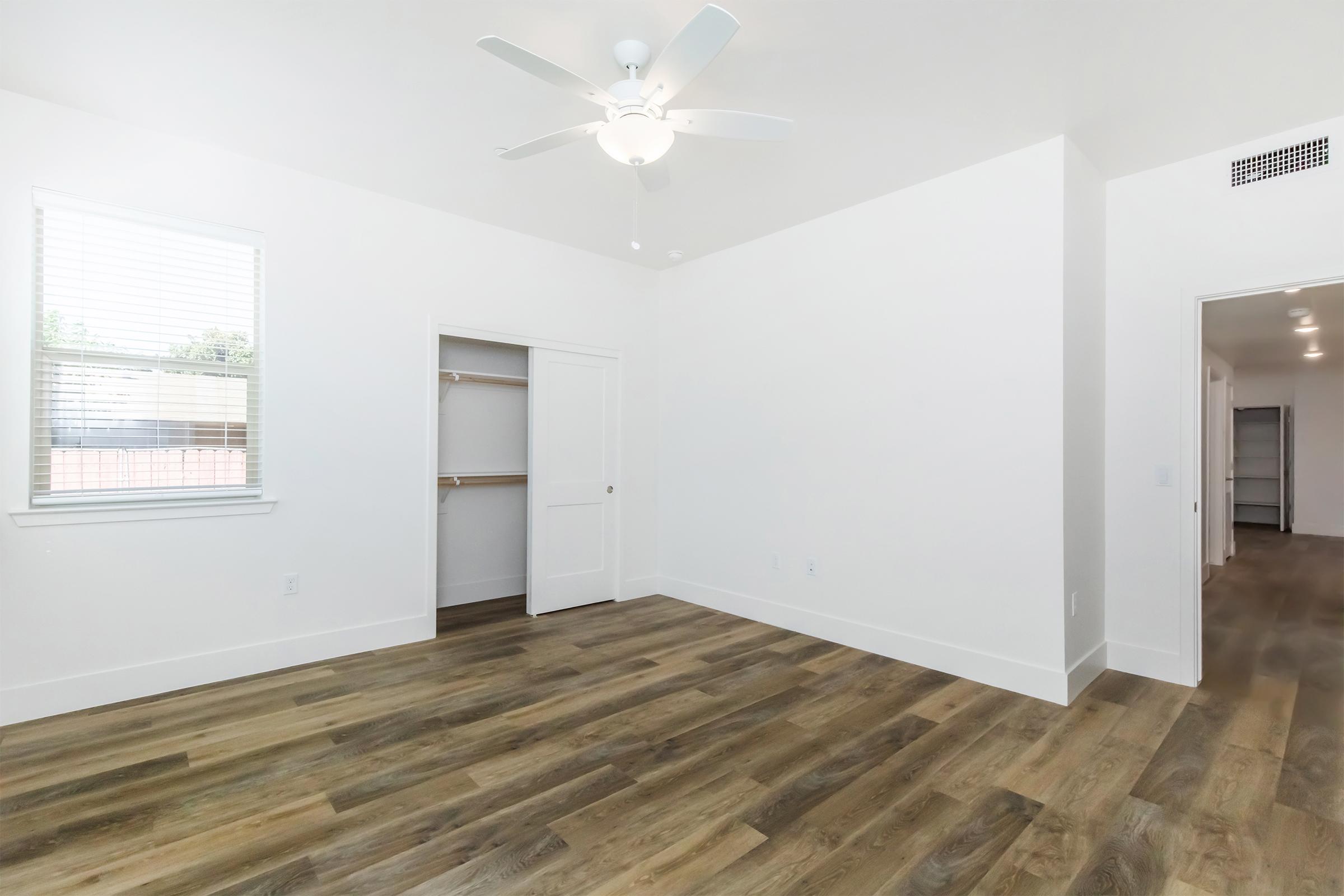 A spacious, empty bedroom featuring light-colored walls, a ceiling fan, and large windows allowing natural light. The room has a closet with an open door, and there is a hallway visible leading to another area. The flooring is wood-like laminate, adding warmth to the modern design.
