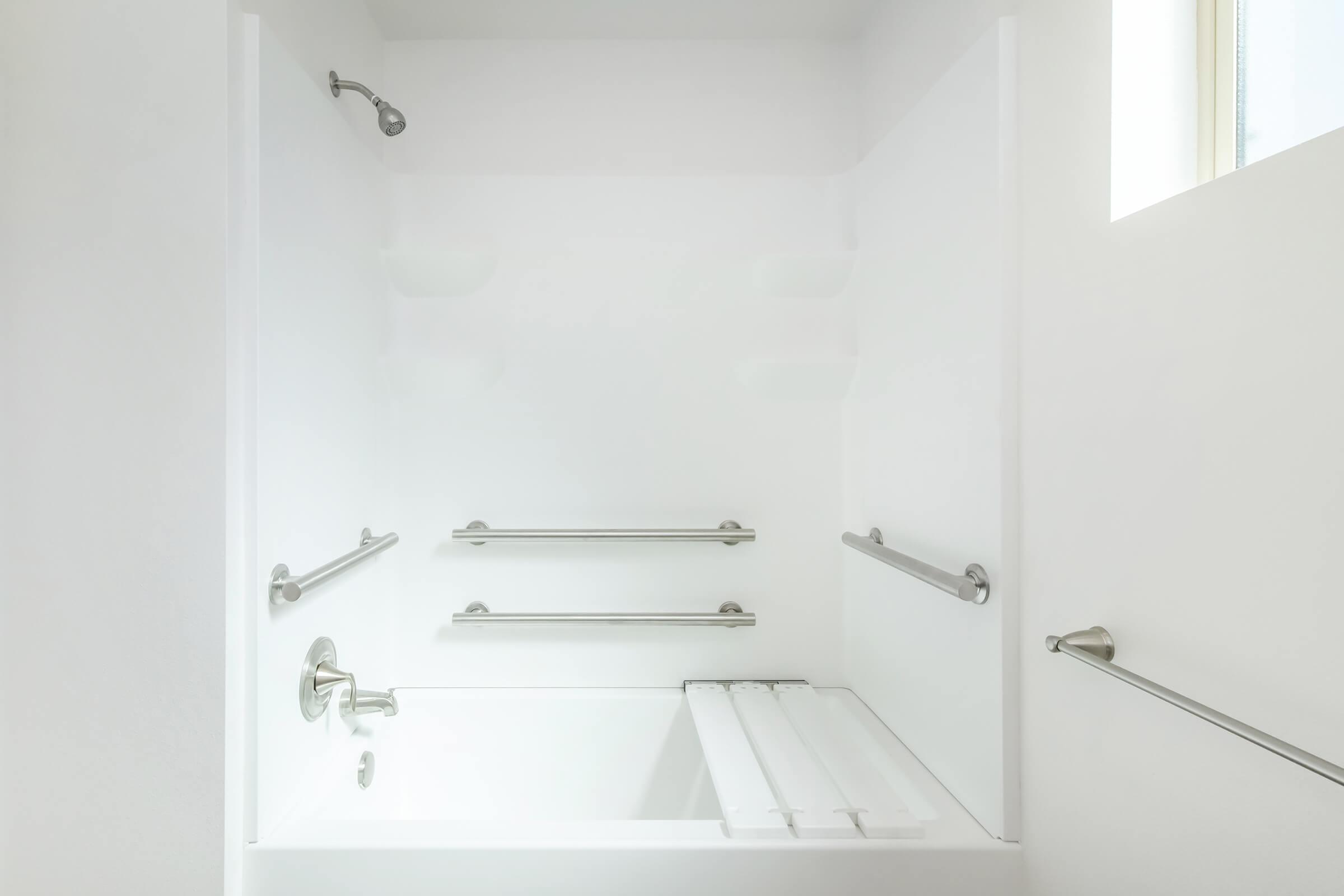 A clean, modern bathroom featuring a white bathtub with a showerhead above, grab bars on the walls for safety, and a small window allowing natural light. The design emphasizes simplicity and accessibility.