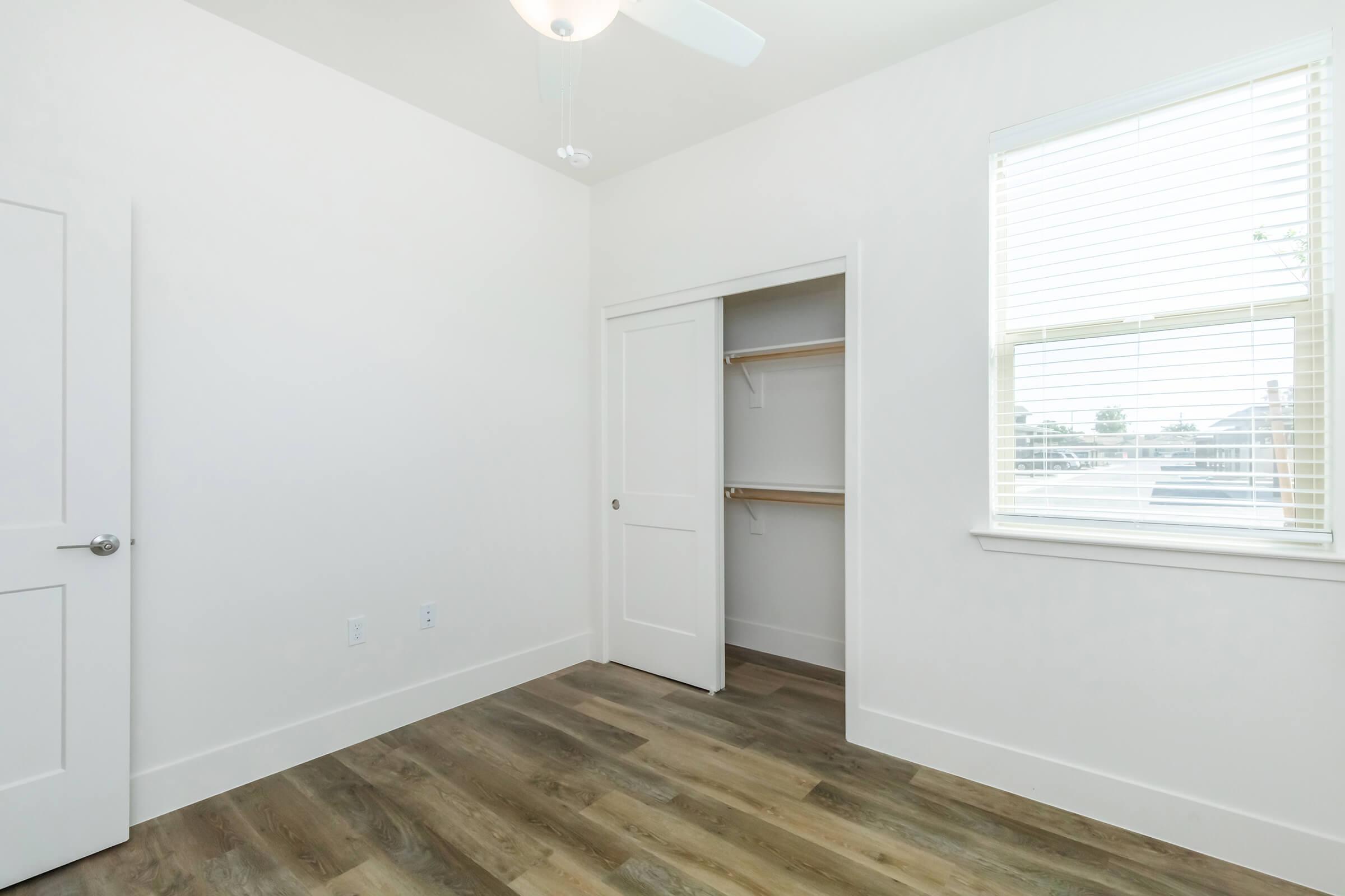 A bright, empty room with white walls and a light wood floor. There's a closed closet with wooden shelves, a window with white blinds allowing natural light, and a ceiling fan. The space is minimalistic and inviting, suitable for various uses or personal decoration.