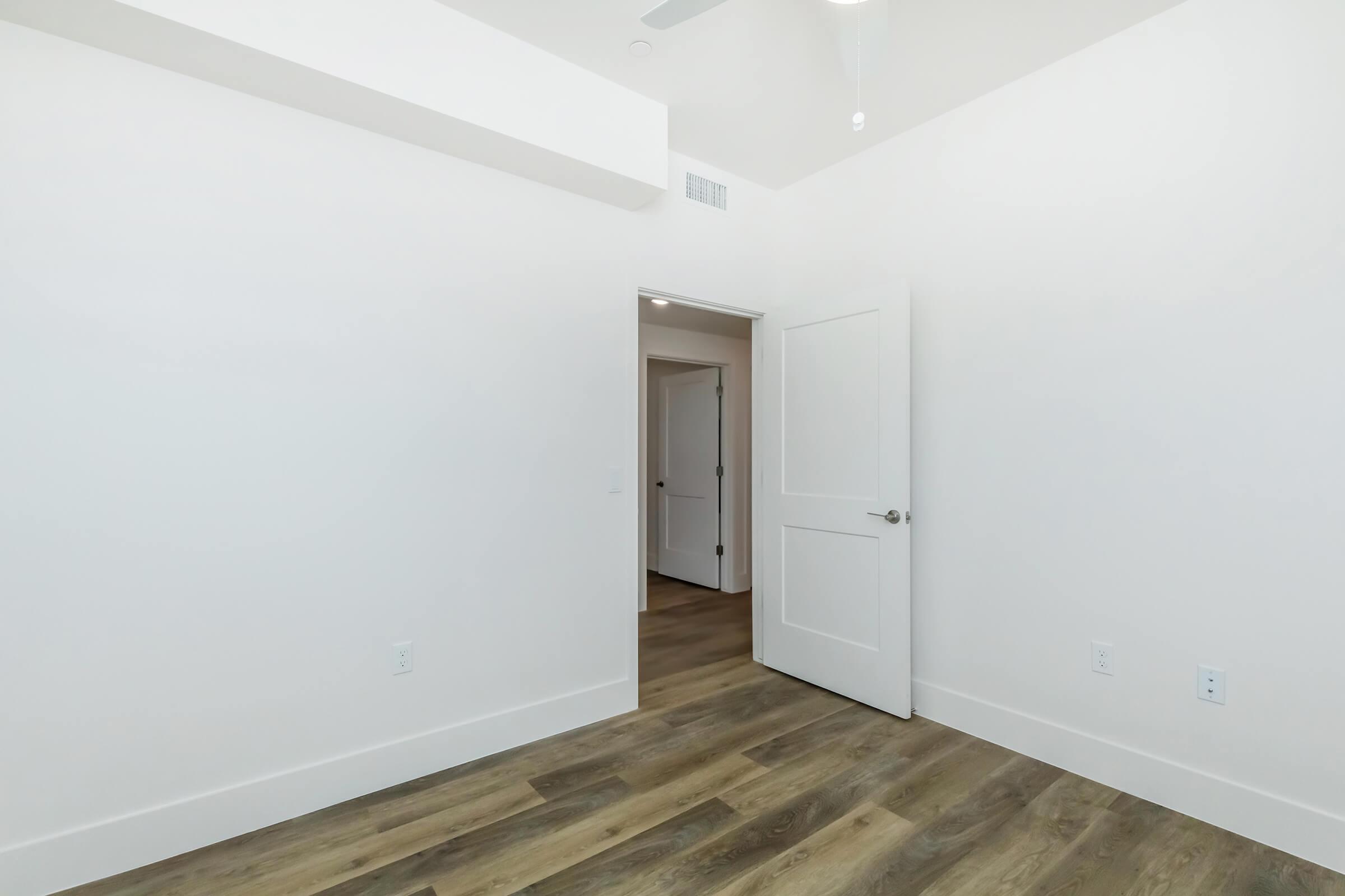 A minimalist room with white walls and a light wooden floor. There's an open door leading to another room, and the space is well-lit with natural light. The ceiling features a ceiling fan, and the overall appearance is clean and modern.