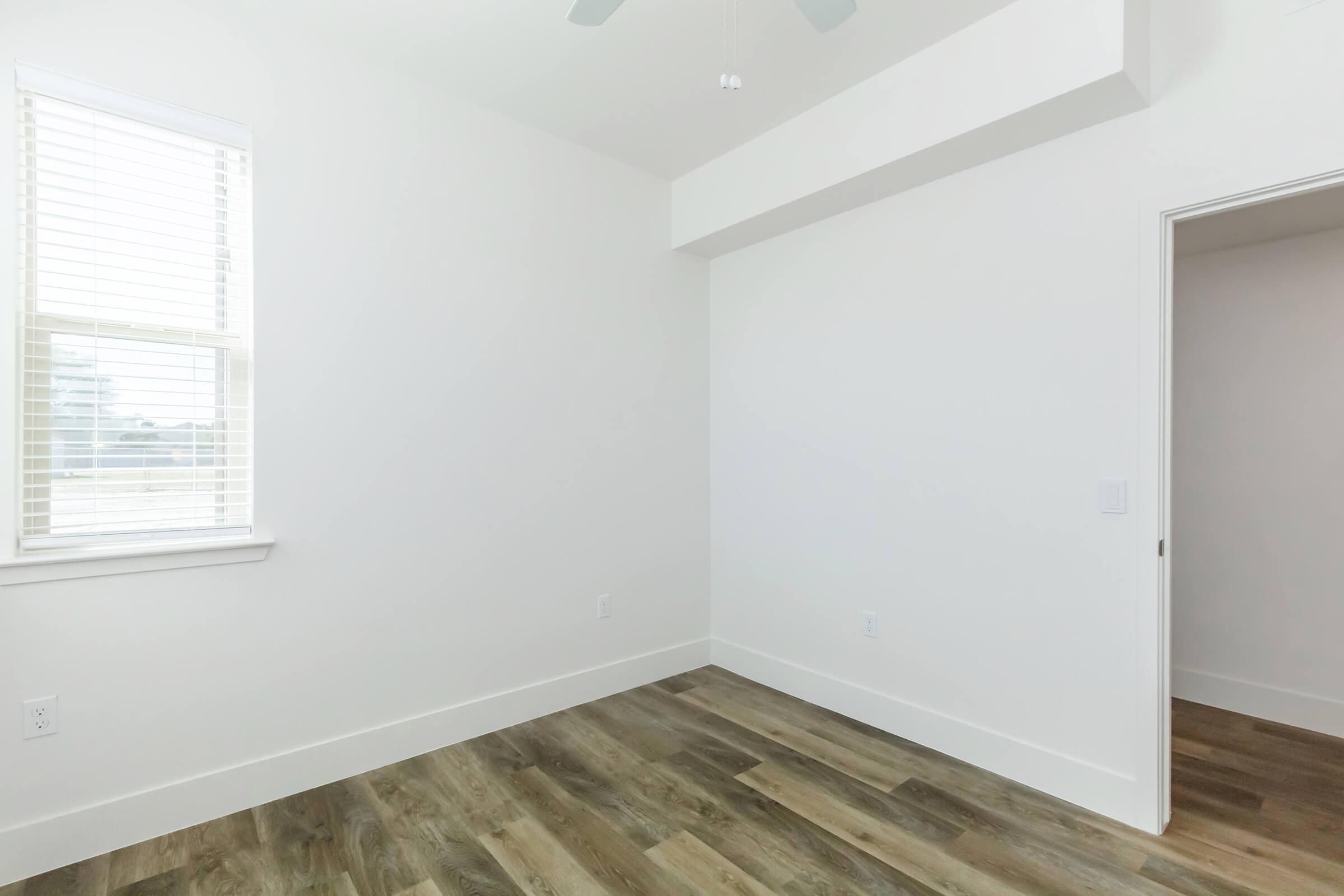 A spacious, empty room with white walls, a ceiling fan, and natural light streaming through a window with blinds. The room features a light wood floor and a door leading to another space, creating a clean and minimalistic aesthetic.
