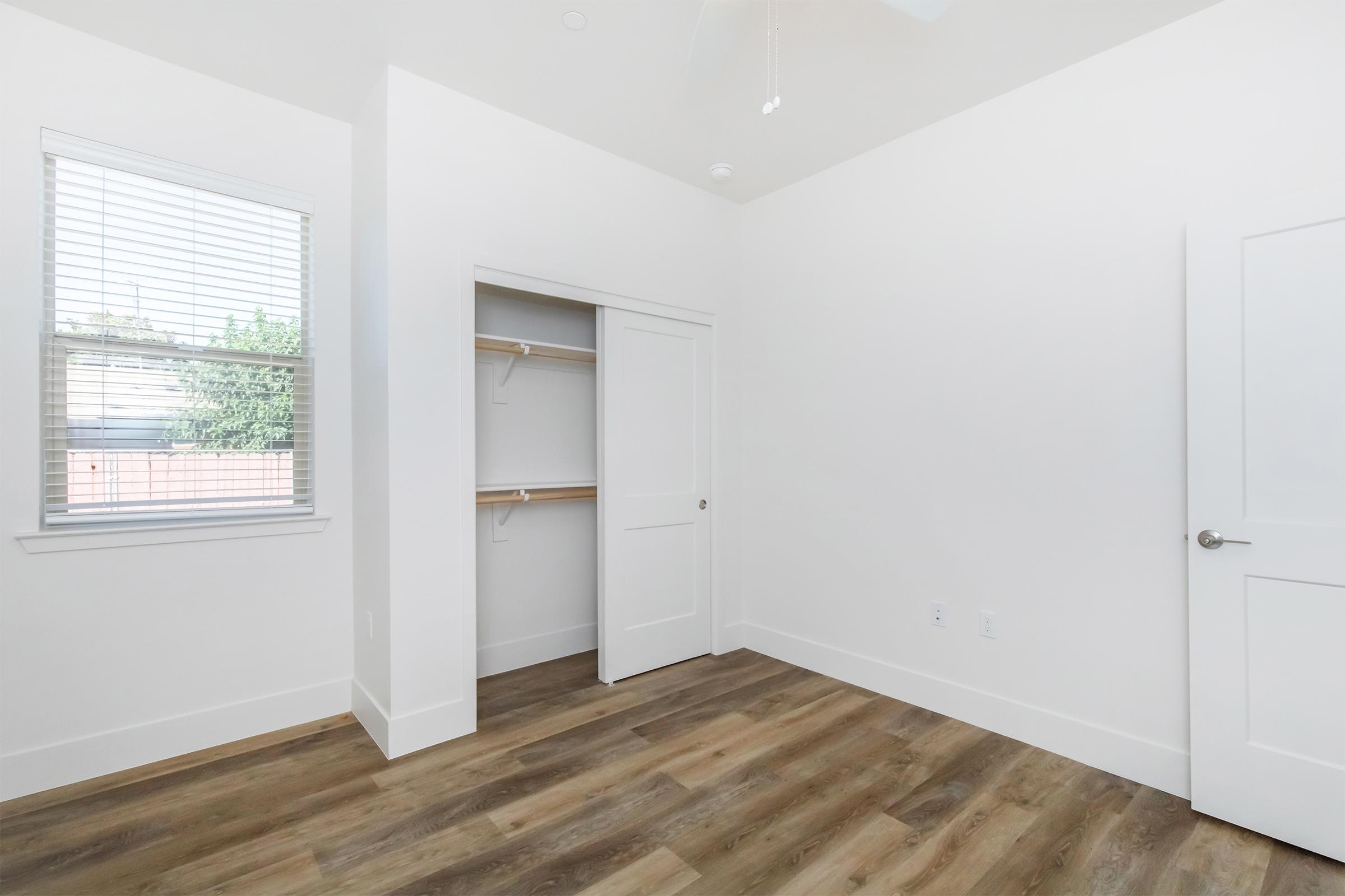 Empty room with light-colored walls, wooden flooring, a ceiling fan, and a closet with sliding doors. A window allows natural light to enter, and there are shelves inside the closet. The overall aesthetic is modern and minimalistic.