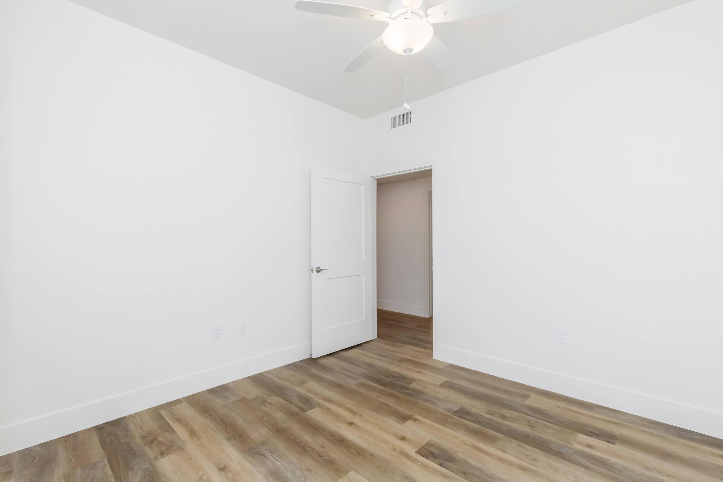 A spacious, empty room with light-colored walls and a wooden floor. A ceiling fan is installed, and there is an open white door leading to another area. The overall aesthetic is clean and modern with ample natural light.