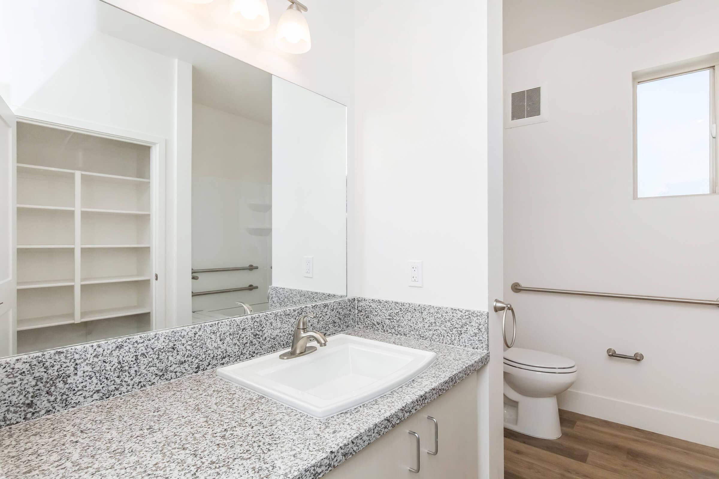 A modern bathroom featuring a large mirror above a granite countertop with a white sink, a toilet in a separate nook, and a window letting in natural light. The space includes a grab bar and shelving in the background, creating a clean and functional design.