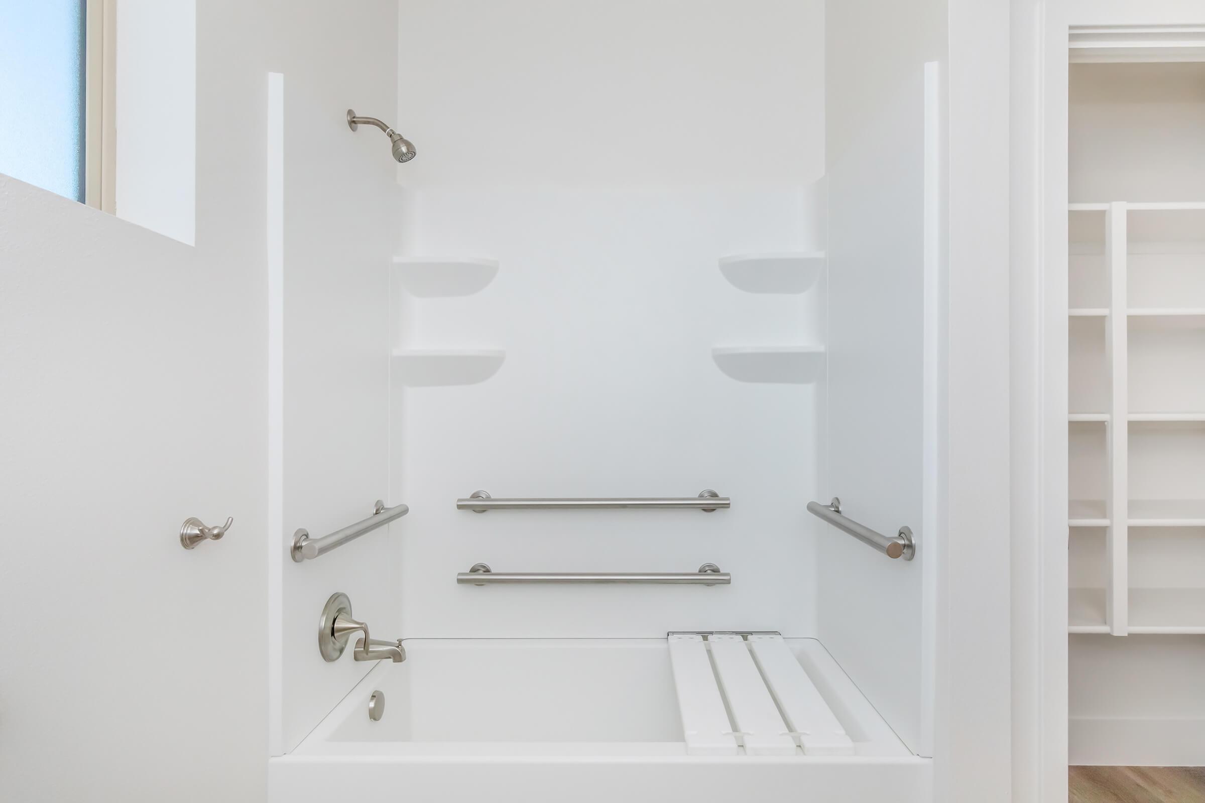 A clean, modern bathroom featuring a white bathtub with safety grab bars, built-in shelves, and a showerhead. The walls are plain and bright, enhancing the spacious feel of the area. Natural light comes in through a window, and there is a storage closet visible in the background.