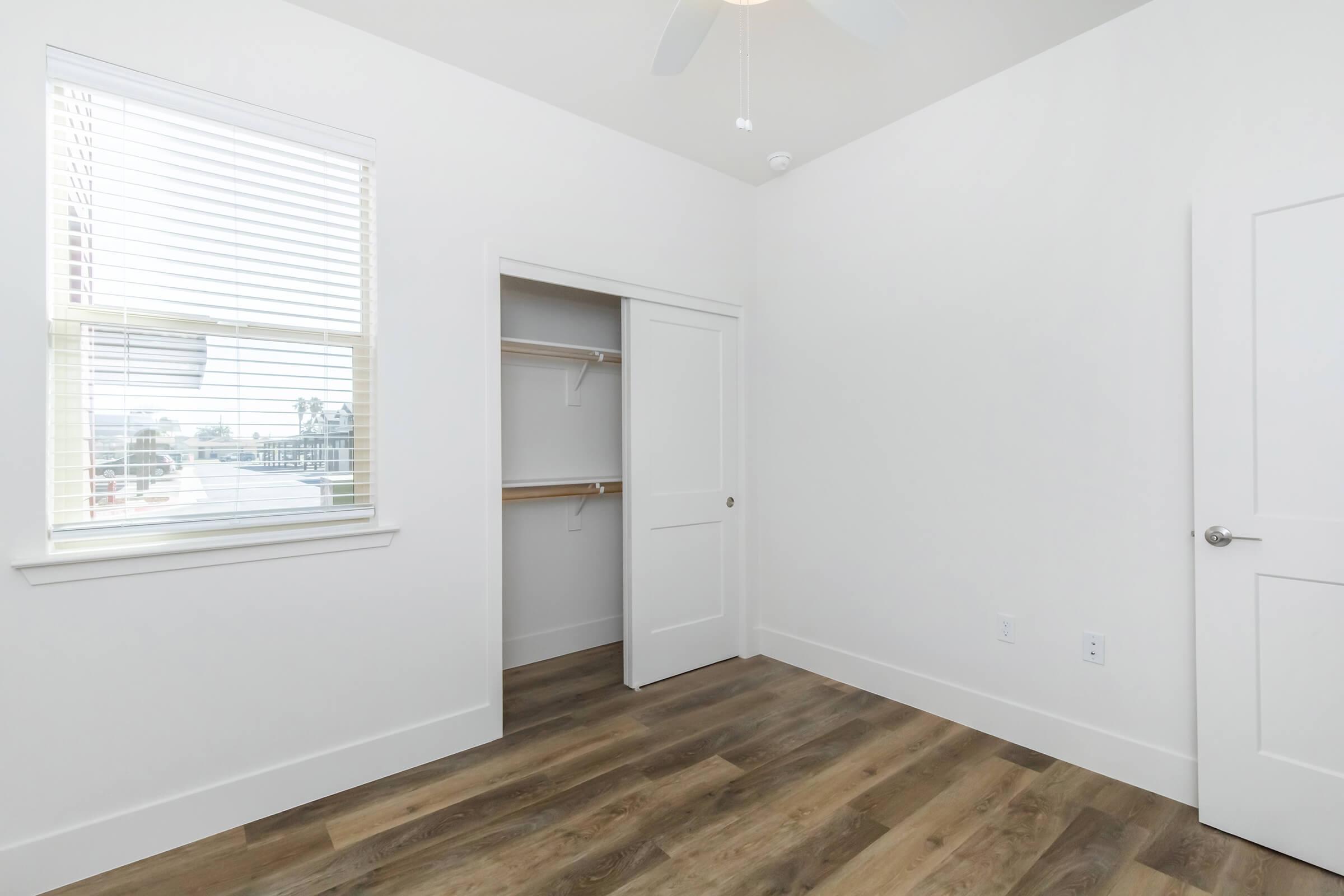 Empty room with white walls, a ceiling fan, and hardwood flooring. A window features blinds, allowing natural light. There's a closet with shelves to the left and a closed door on the right. The space appears bright and modern, suitable for furniture placement.