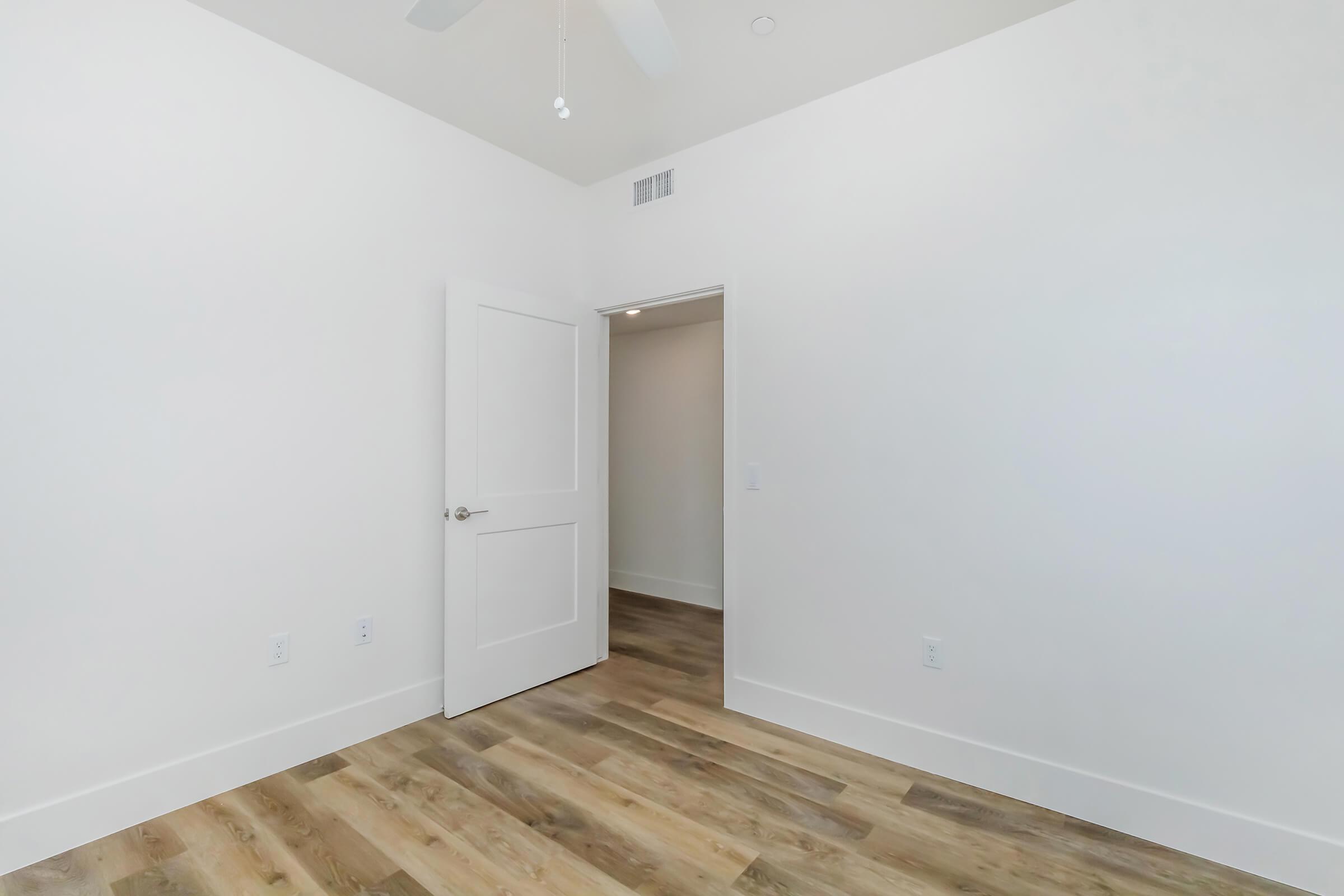 A vacant room with light-colored walls, featuring a wooden floor. A door is visible on the left side leading to another space, while a ceiling fan is mounted above. The room has a minimalist design and ample natural light.