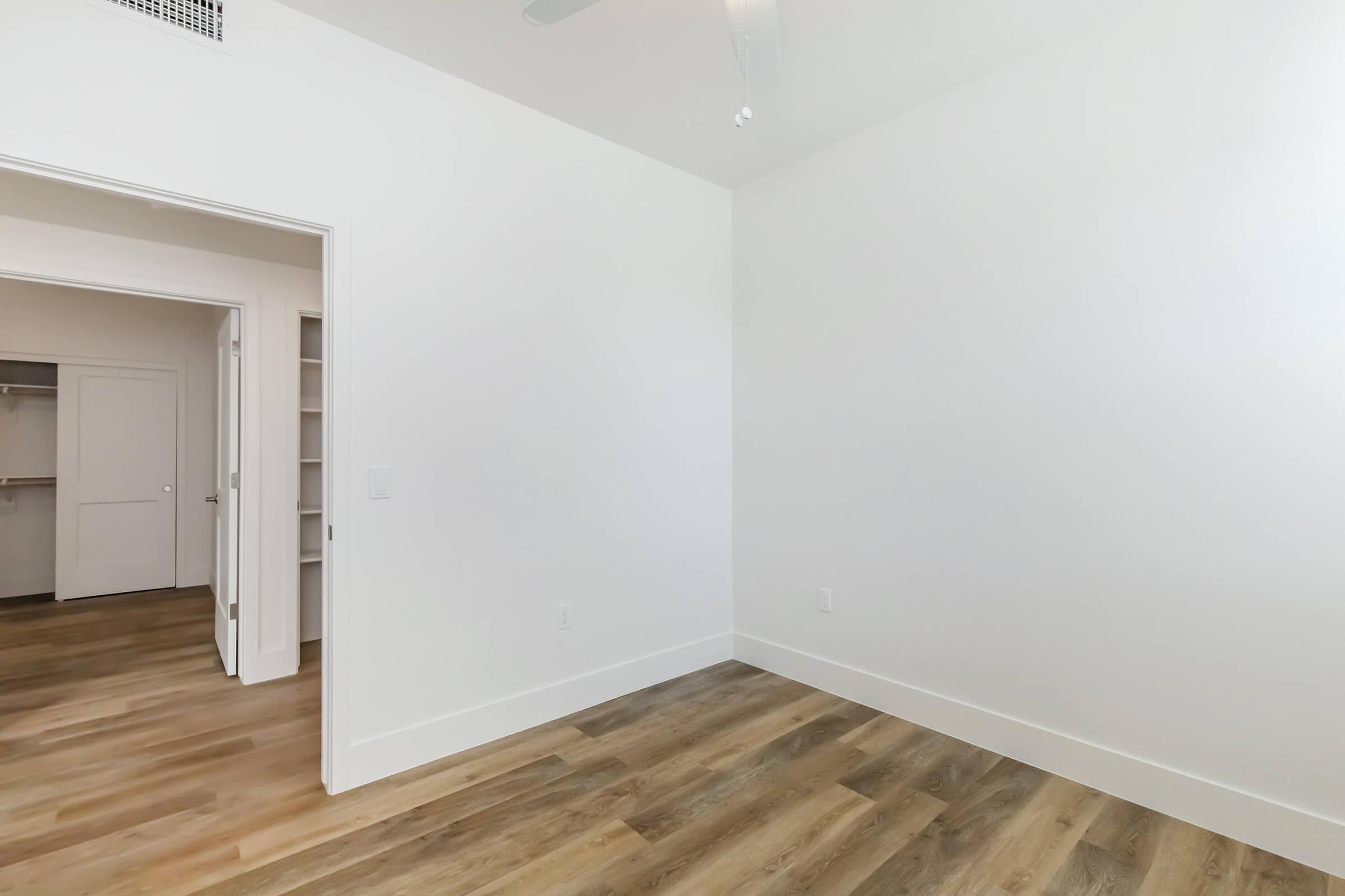 Empty interior space with white walls and light brown wooden flooring. A doorway leads to another room with a closet visible. The ceiling has a ceiling fan, and the overall ambiance is bright and minimalist.