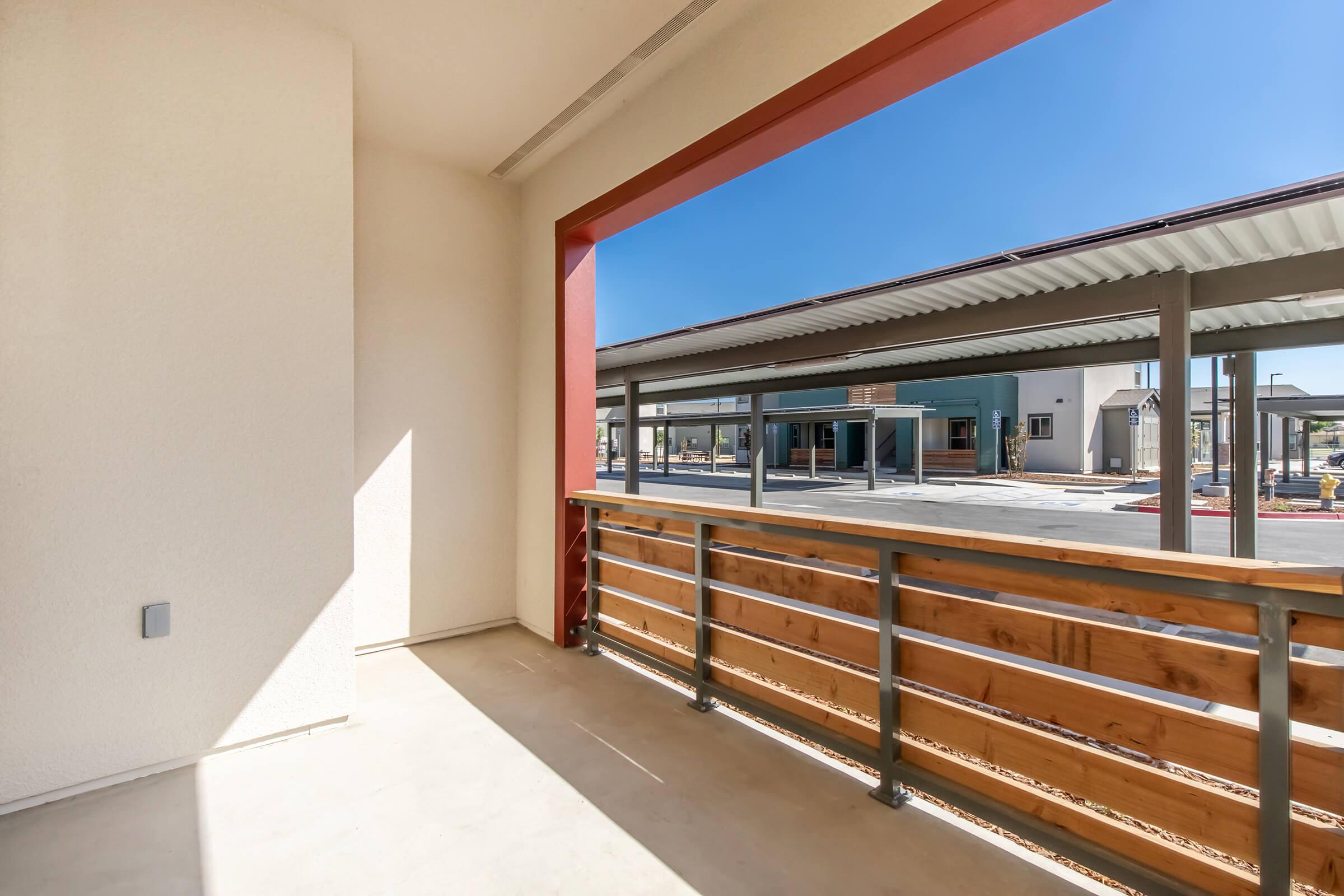 A view from a balcony showing a partially covered outdoor area with wooden railing. In the background, there are buildings with flat roofs and a clear blue sky overhead. The space appears modern and well-lit.