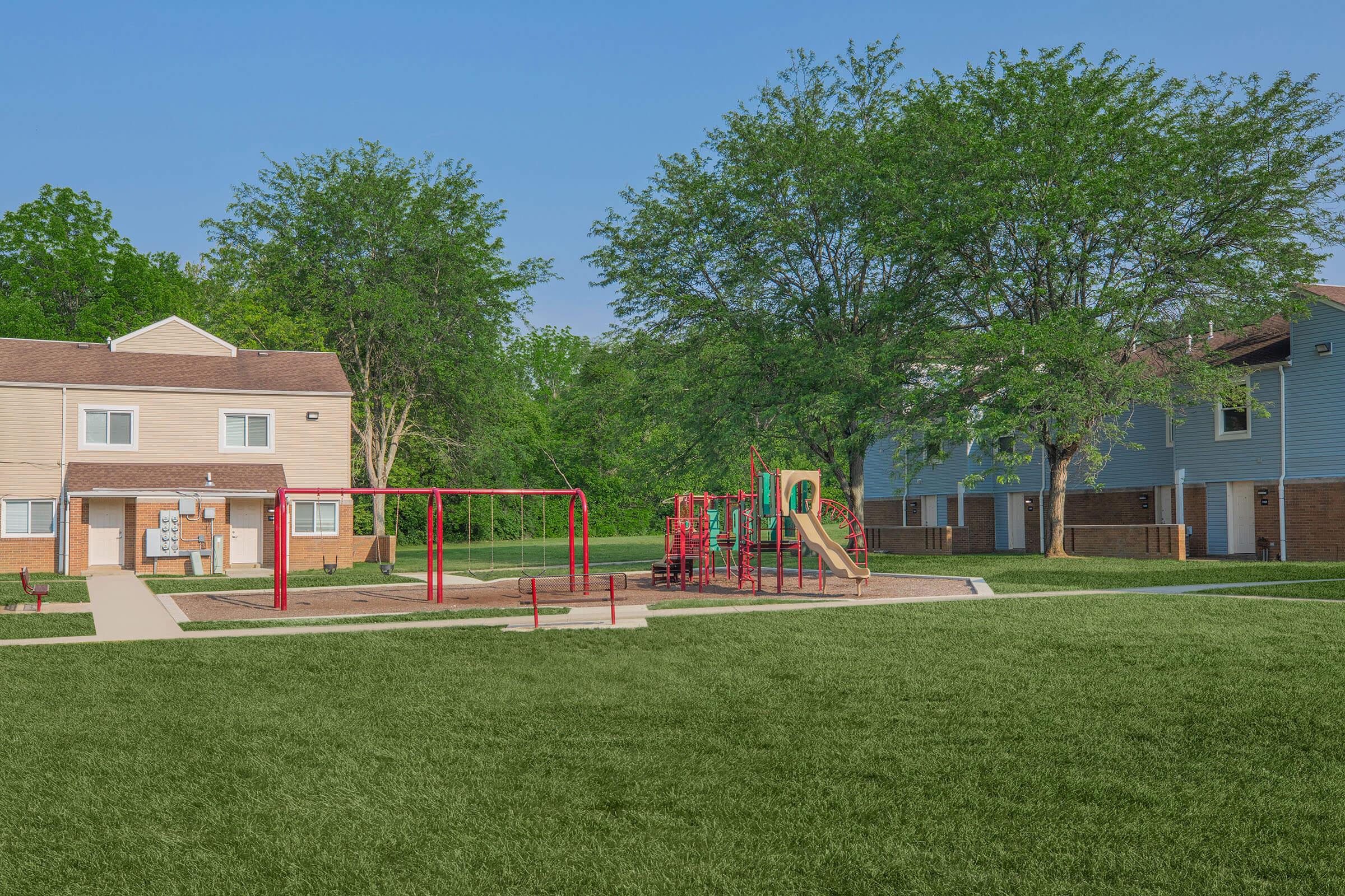 A playground with a slide and swings surrounded by green grass, located in a residential area. There are two multistory buildings in the background, and trees providing shade. The scene is bright and sunny, indicating a clear day.