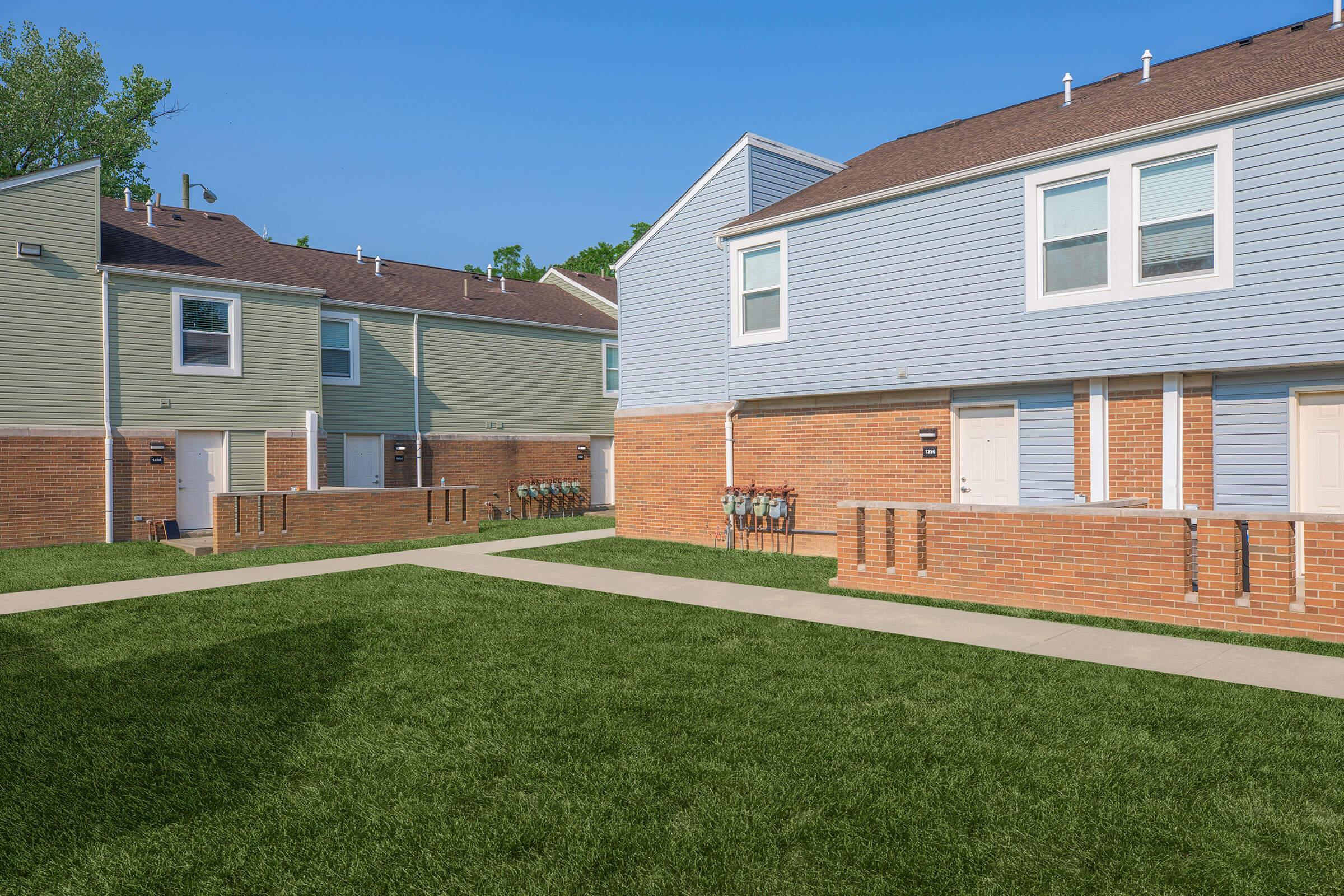 A view of a residential area featuring two multi-family homes with pastel-colored siding, surrounded by well-maintained green lawns. Walkways connect the buildings, and utility meters are visible along one side of the houses. The sky is clear and blue, giving a bright and welcoming atmosphere.