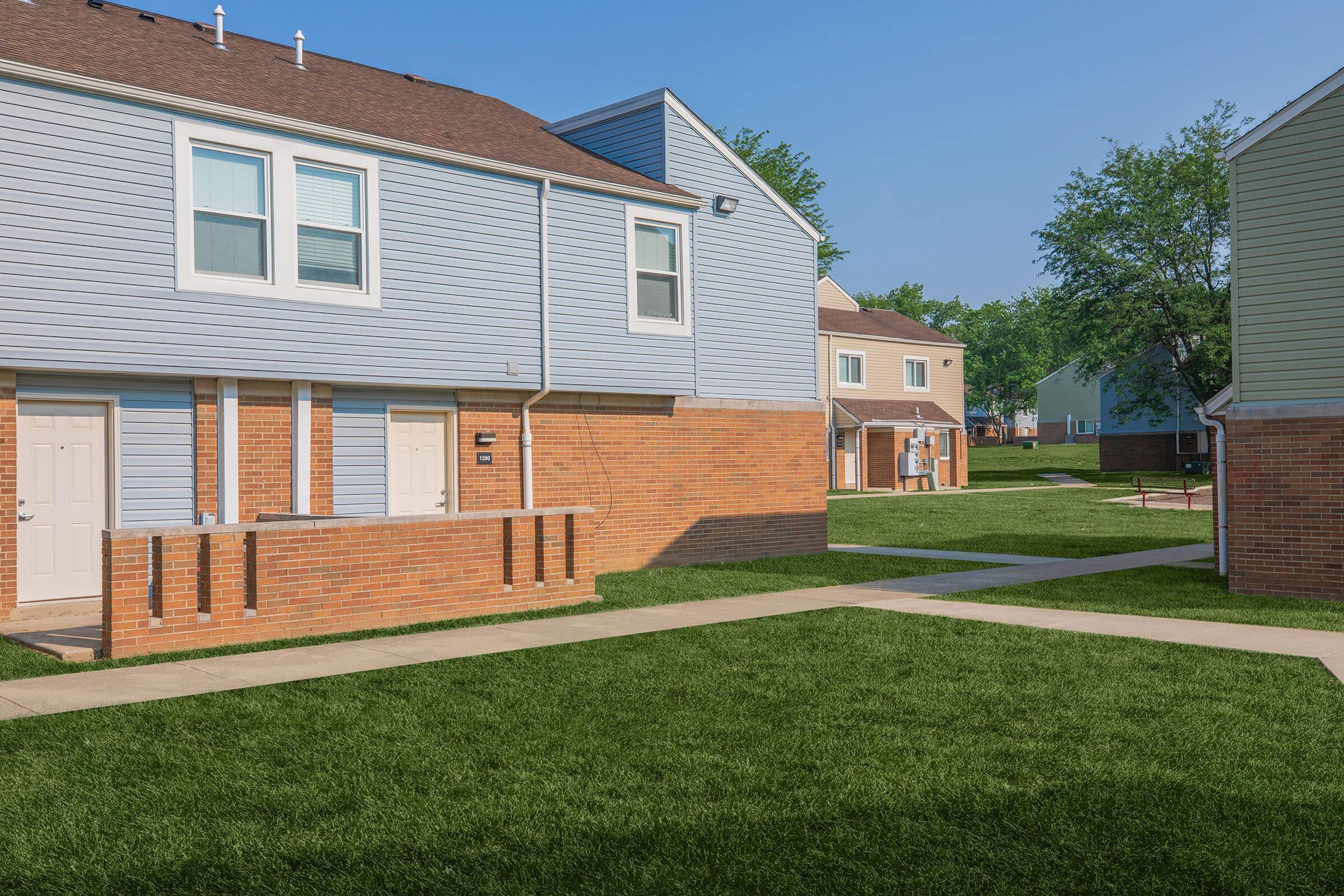 A view of a residential area featuring several two-story townhouses with blue and beige siding. The houses are separated by grassy lawns and sidewalks, under a clear blue sky. Trees are visible in the background, adding to the neighborhood ambiance.