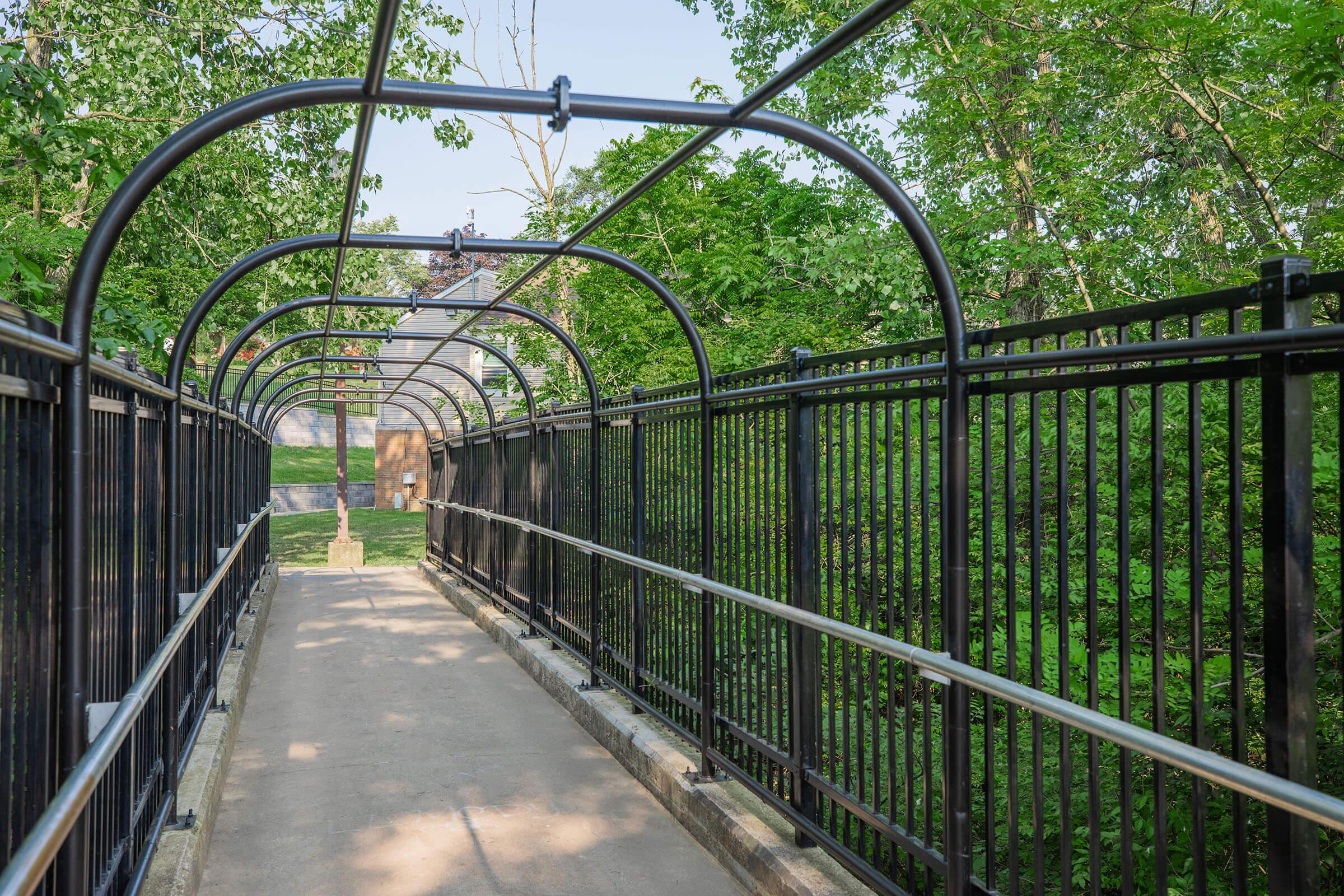 A winding pathway enclosed by a black metal arch structure, flanked by greenery on both sides. The walkway leads towards a small wooden building in the distance, surrounded by trees, creating a peaceful, nature-inspired atmosphere.