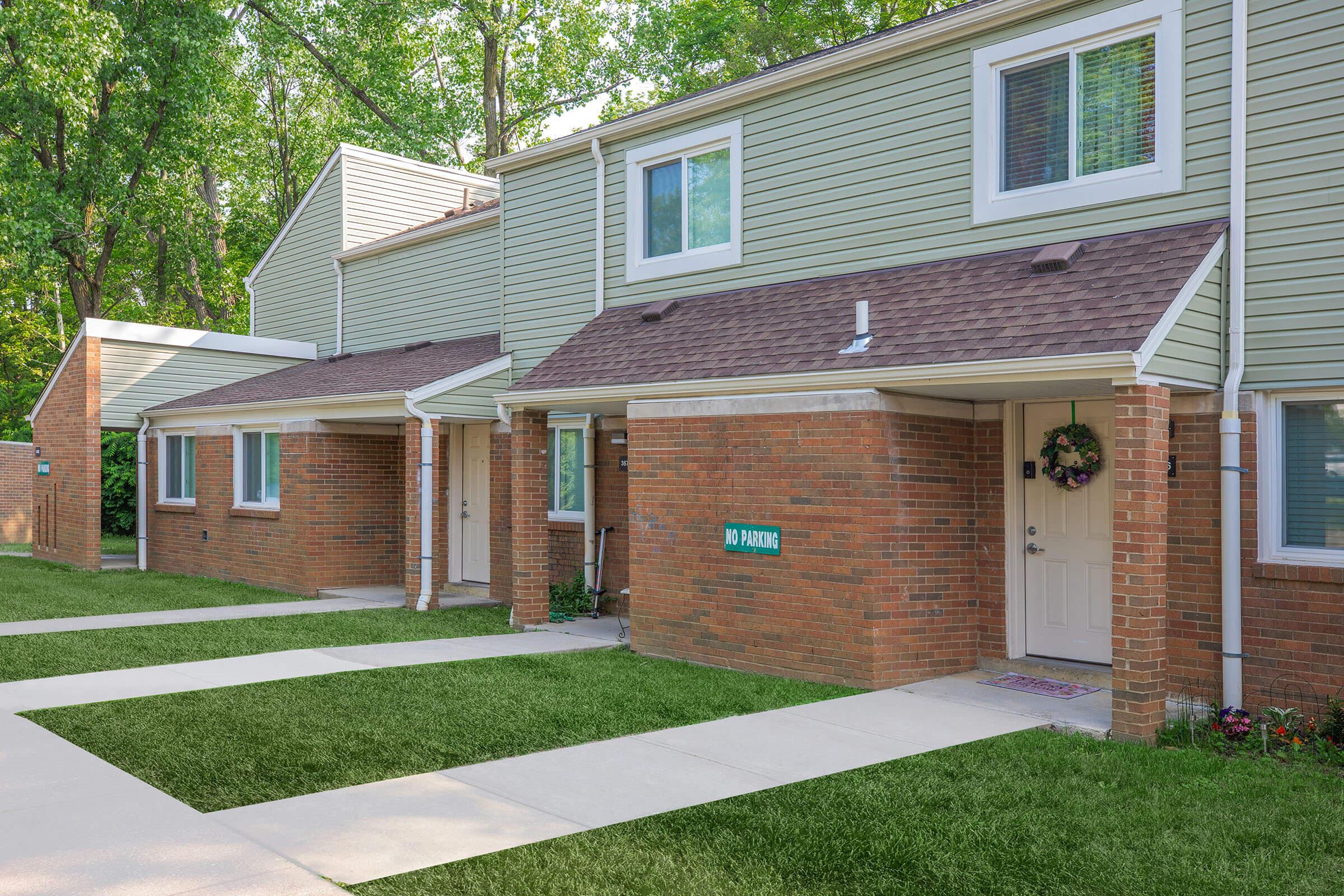 Row of townhouses with green siding and brown roofs. Each unit has a front door with a small stoop and a covered porch area. The pathway is made of concrete, leading to neat, green grass in front of the homes. Small trees and greenery line the background, creating a vibrant, welcoming atmosphere.