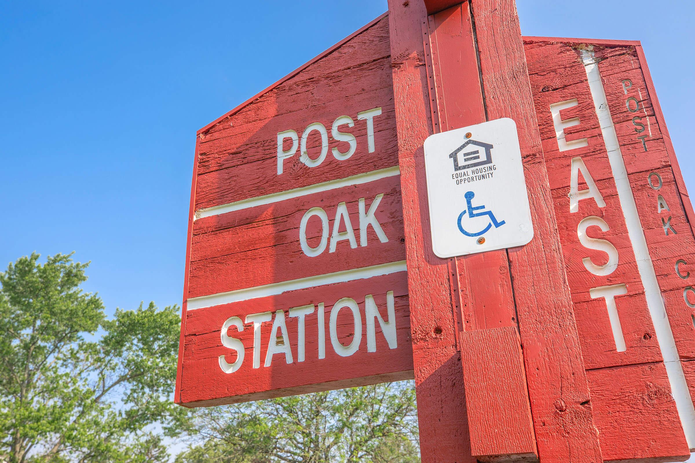 A weathered red sign indicating "Post Oak Station" with directional information pointing east. There is a white sign below it displaying the symbol for accessible facilities, promoting equal housing opportunity. Surrounding greenery and a clear blue sky are visible in the background.