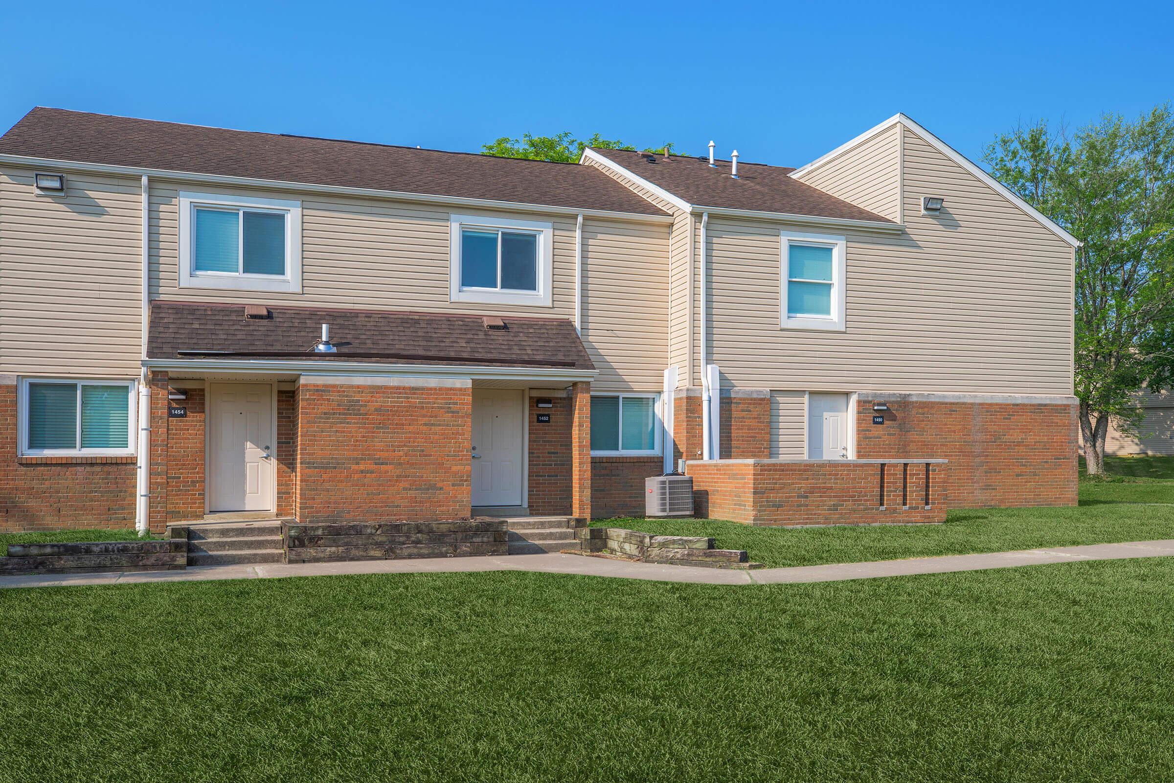 Two-story residential building featuring a combination of brick and siding. The front has two separate entrances, with small steps leading to each door. There is a grassy area in front, and a clear blue sky is visible overhead, enhancing the bright and open ambiance of the setting.