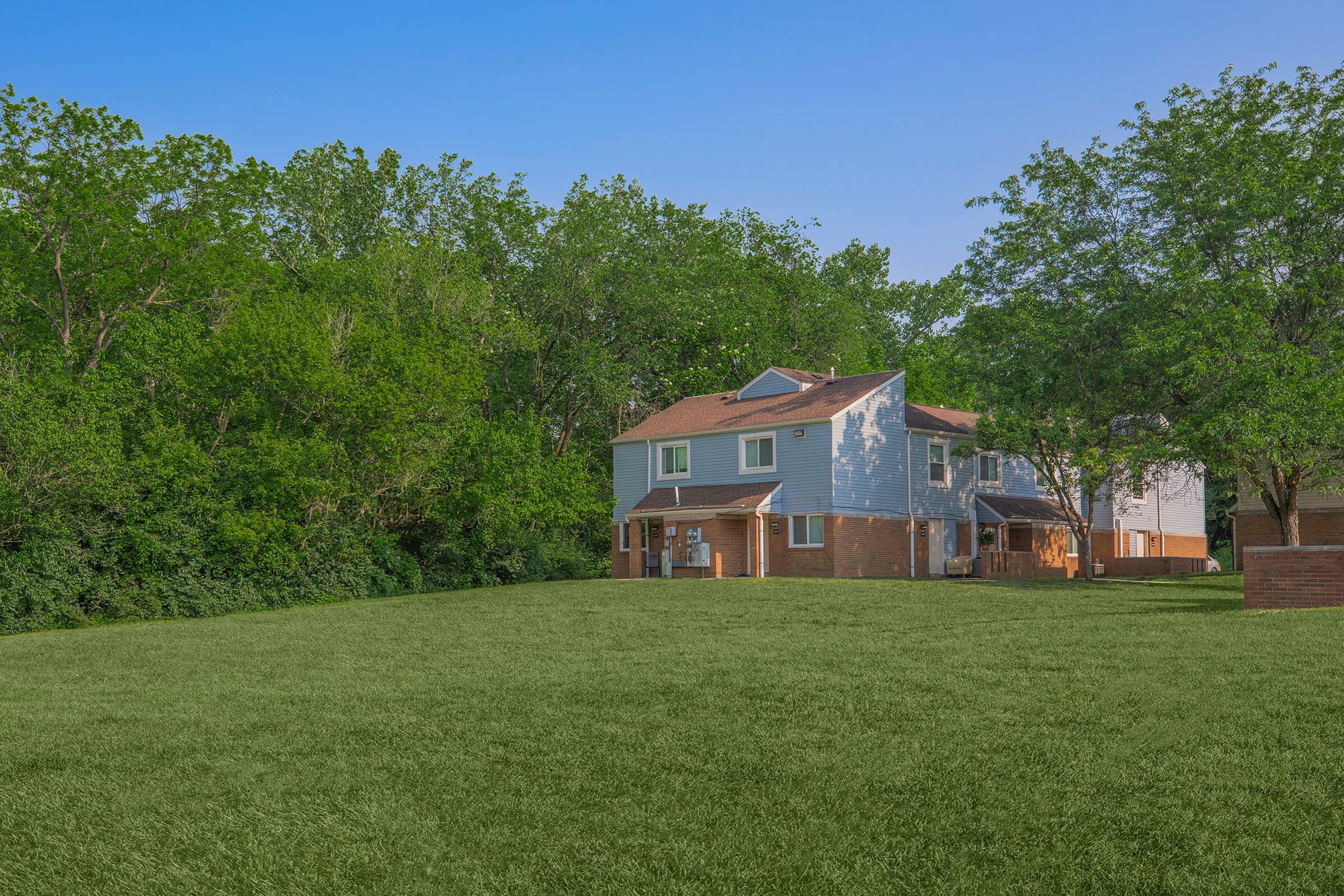 Two multi-story residential buildings with light blue and brick exteriors, nestled on a green lawn. Behind the buildings, a dense row of trees provides a natural backdrop against a clear blue sky. The scene conveys a tranquil suburban environment.