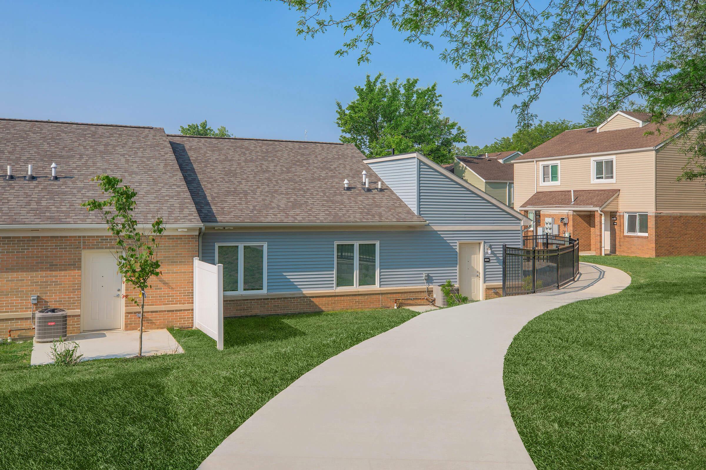A pathway winding through a residential area with two multi-unit buildings. One building features a brick facade, while the other has a light blue exterior. Lush green grass surrounds the walkway, and trees provide greenery in the background under a clear blue sky.