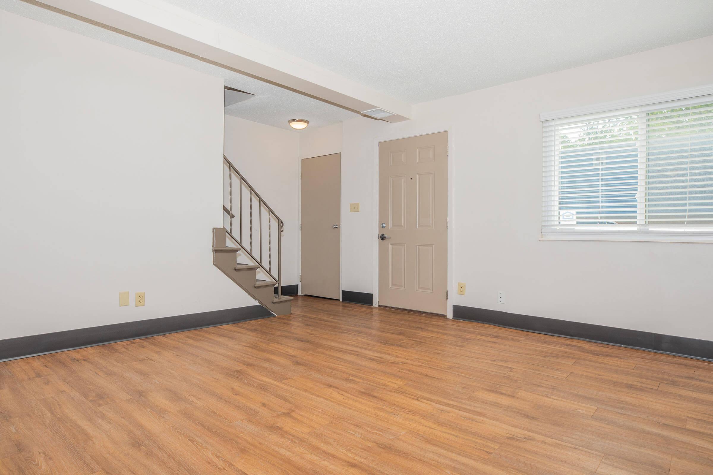 A vacant room with wooden laminate flooring, featuring a staircase leading to an upper level. There is a closed door on the right and a window with blinds allowing natural light in. The walls are painted light-colored, creating a bright and open atmosphere.