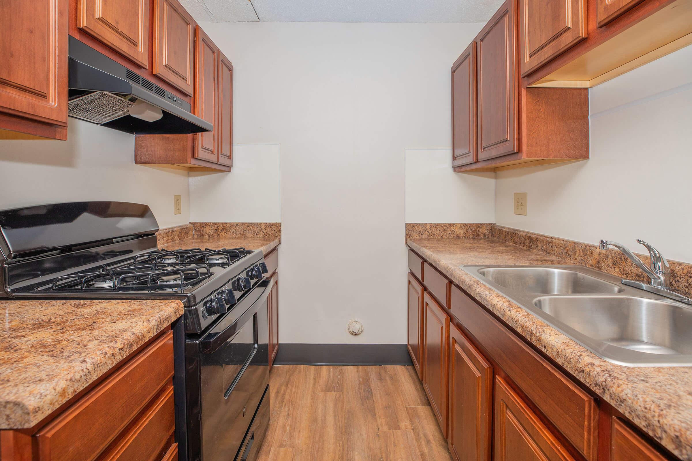 A view of an apartment kitchen featuring dark wooden cabinets, a black gas stove, and a double sink with chrome faucets. The countertop is made of a brown granite-like material, and the floors have a wood-like finish. The walls are painted in a light color, creating a spacious feel.