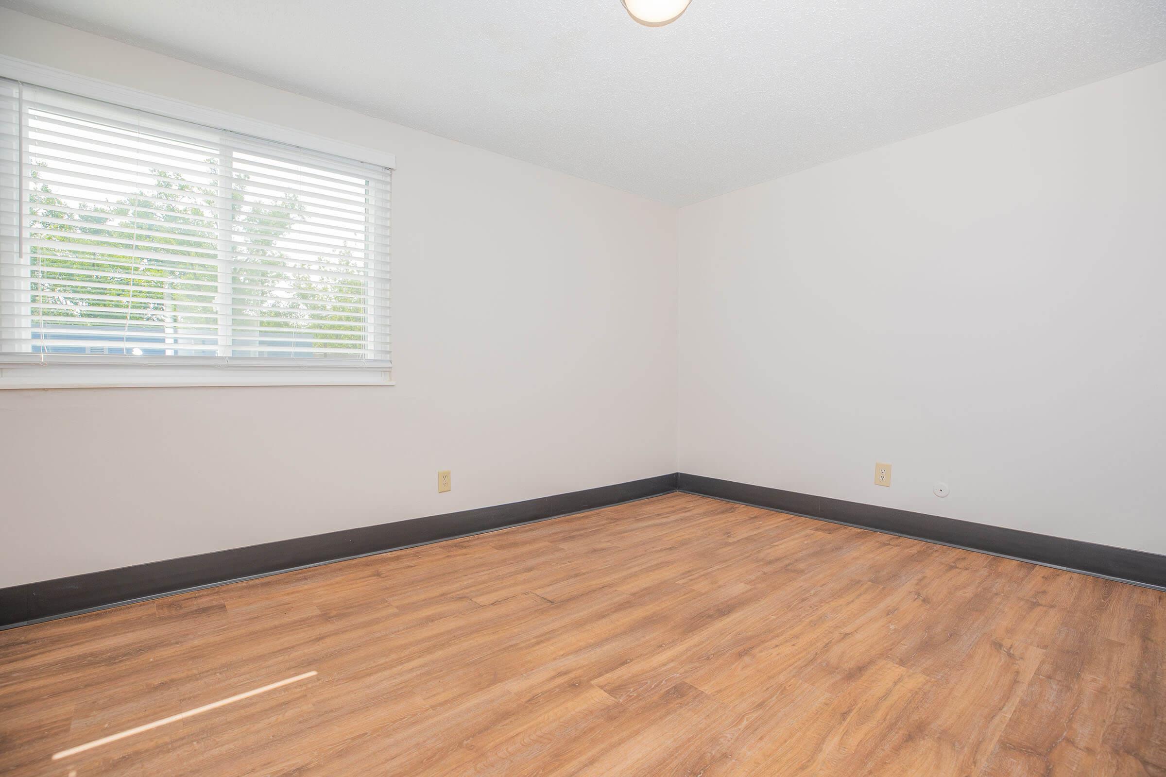Empty room with light-colored walls, a window with white blinds letting in natural light, and a wooden floor. The space is clear of furniture, creating a spacious and minimalist appearance.