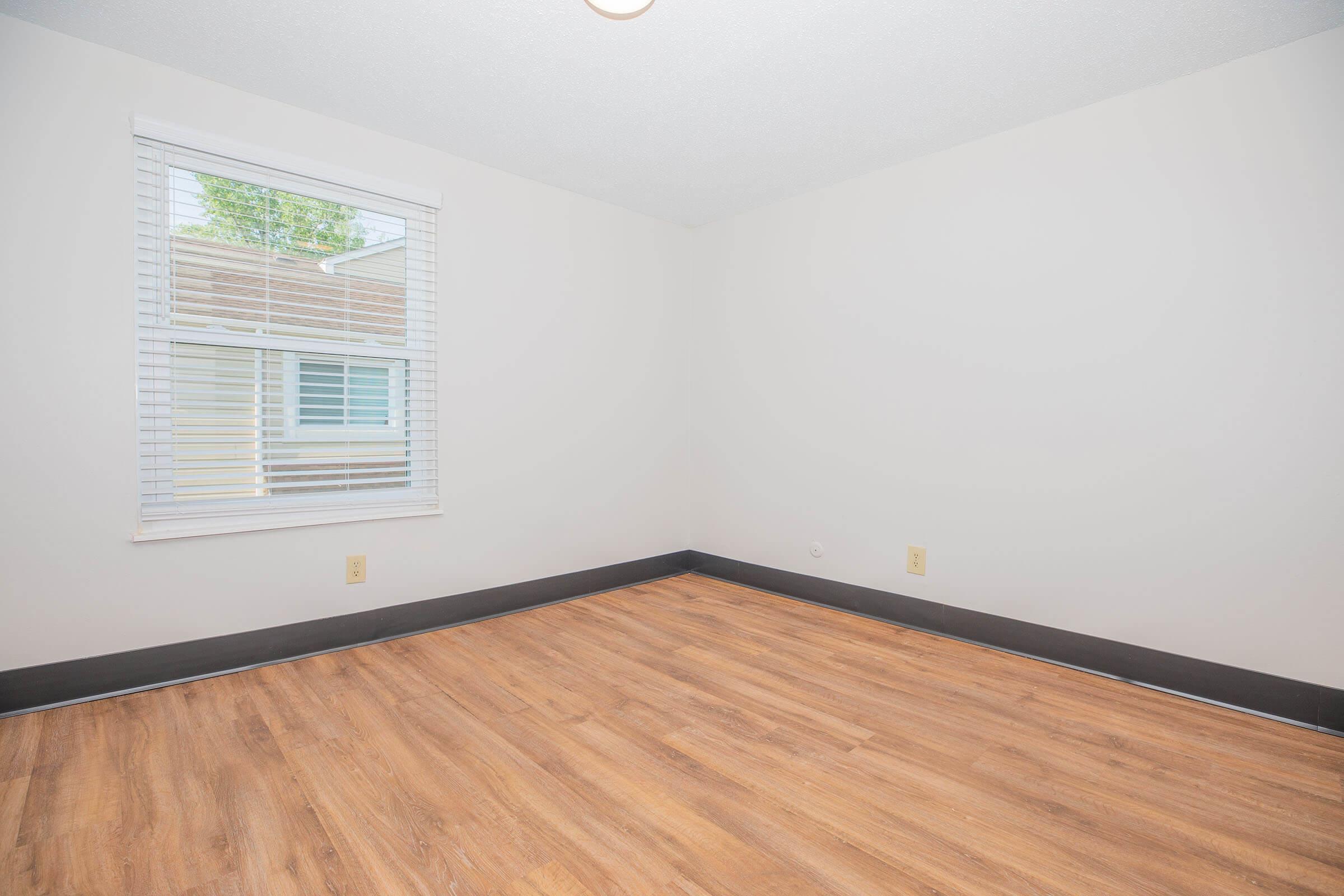 Empty room with light-colored walls and a window with white blinds. The floor has wooden planks, and there are no furnishings present. The room appears bright and spacious, with a simple overhead light fixture.
