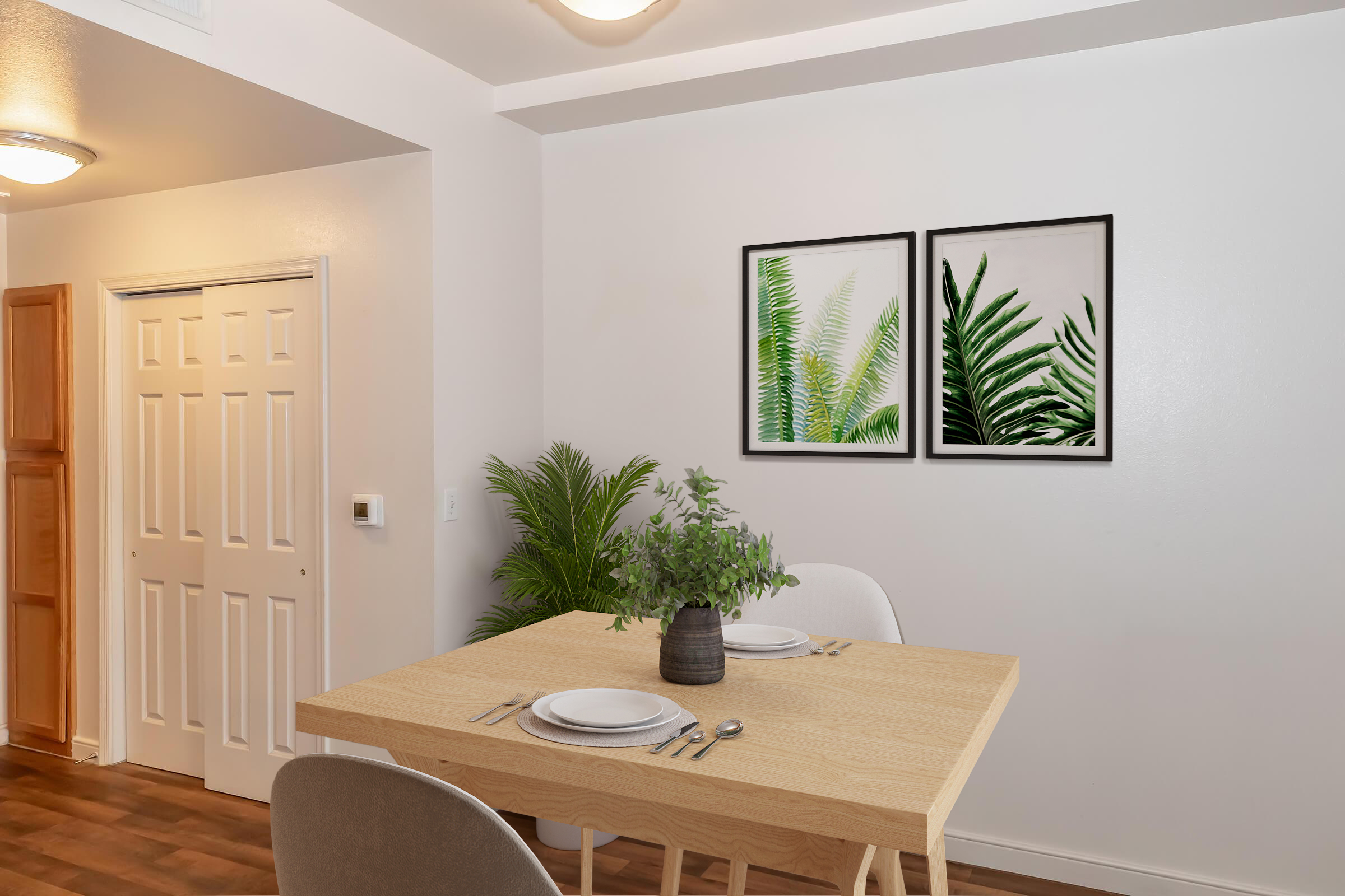A modern dining area featuring a light wood table set for two with simple white plates and utensils. The wall showcases two framed botanical prints of ferns, and a potted plant adds greenery to the space. A door is visible in the background, enhancing the inviting atmosphere.