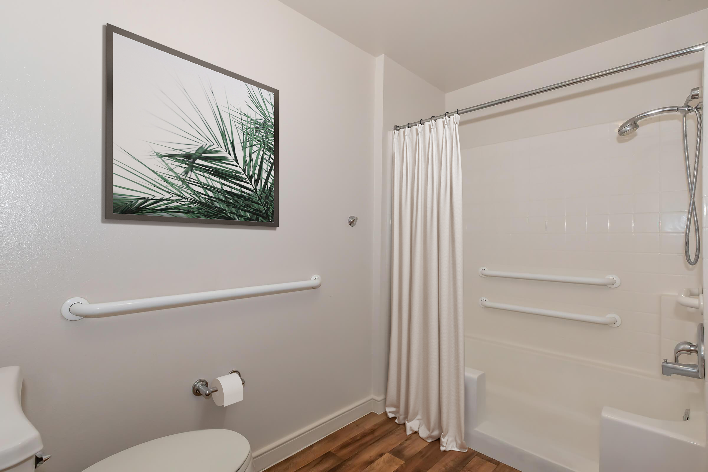 A clean and modern bathroom featuring a white shower with a curtain, a toilet, and safety grab bars. A framed artwork of palm leaves hangs on the wall, enhancing the serene atmosphere. The floor has a wood-like finish, contributing to the contemporary design.