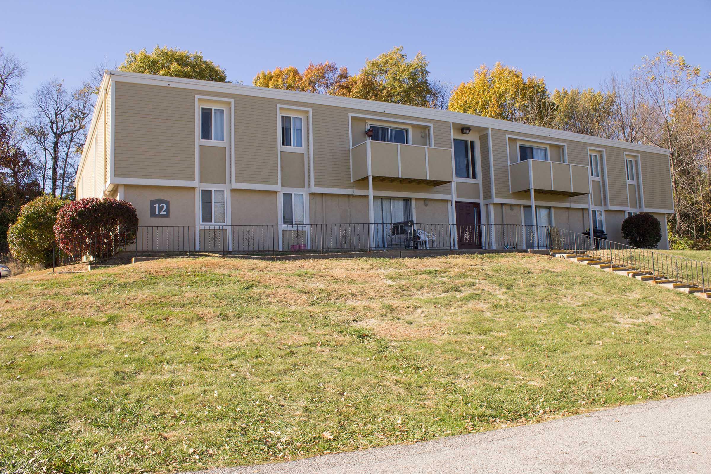 A two-story apartment building with multiple balconies, situated on a grassy hillside. The building features a light-colored exterior and is flanked by trees displaying autumn foliage. A short staircase leads up to the entrance, with a pathway and a number 12 visible on the building.