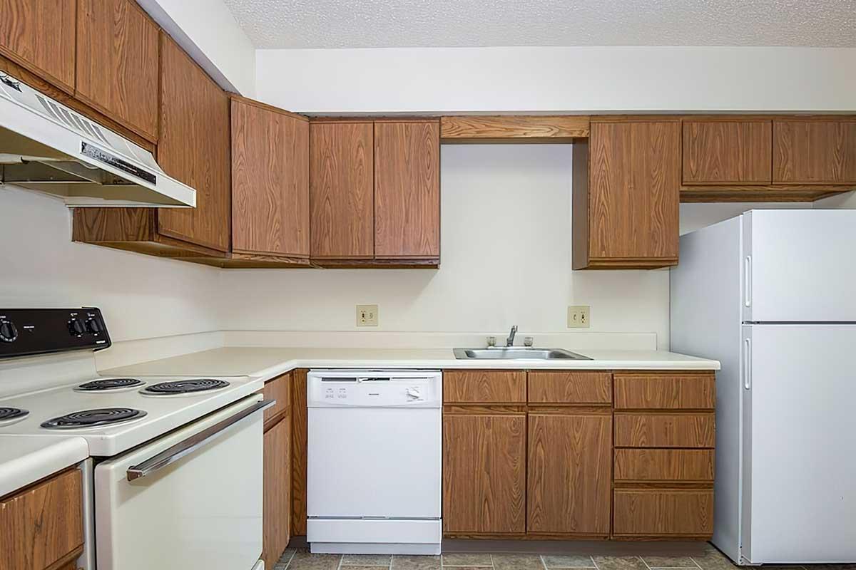 A well-lit kitchen featuring wooden cabinets, a white refrigerator, a dishwasher, a stove with an oven, and a double sink. The countertops are light-colored, and the floor has a textured design. The kitchen appears clean and functional, ideal for cooking and food preparation.