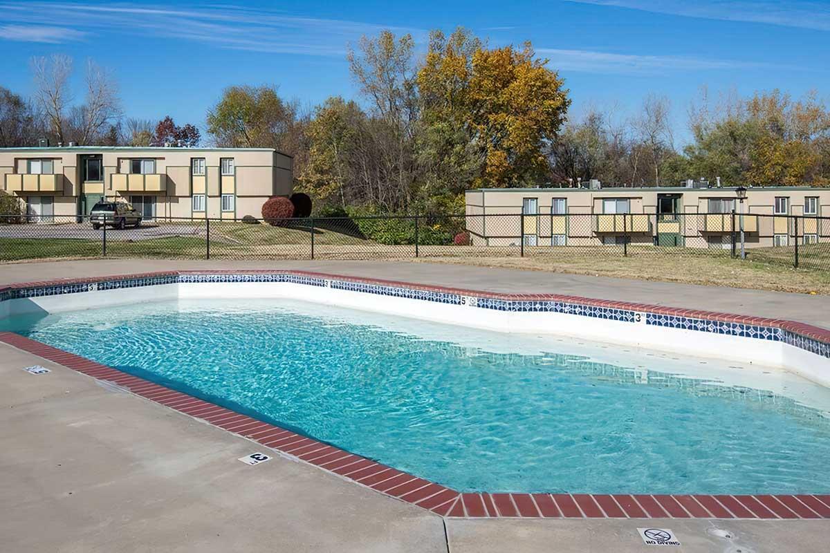 Swimming pool surrounded by a concrete deck, with a fence in the background. Features a few trees with autumn foliage and a row of low-rise buildings. Clear blue sky above, indicating a sunny day.