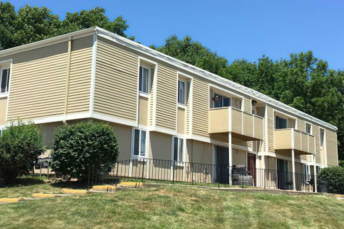 A two-story apartment building with beige siding and multiple windows, surrounded by grass and a few shrubs. The structure features balconies on the upper level and a fenced area in front. Bright blue sky and trees are visible in the background.