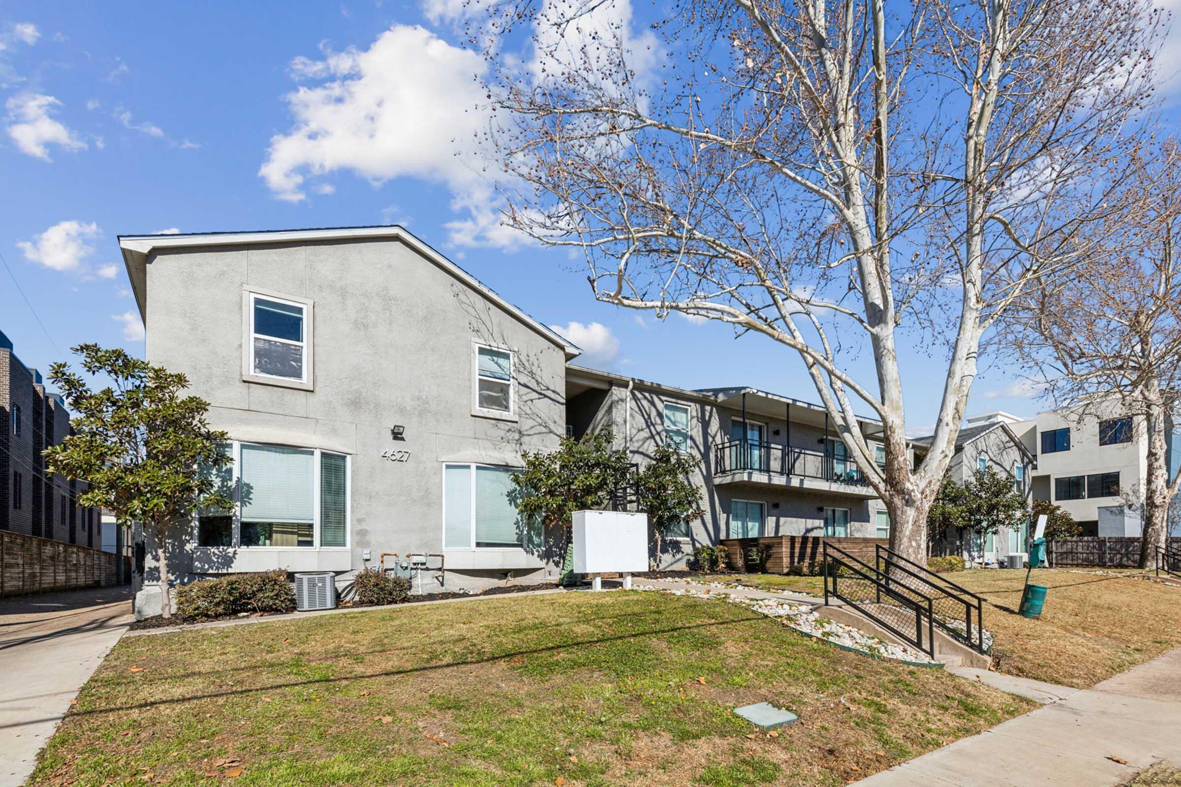A two-story gray apartment building with large windows, surrounded by grass and a few trees. There are easily accessible outdoor walkways and a sign near the entrance. The sky is partly cloudy, creating a bright atmosphere.