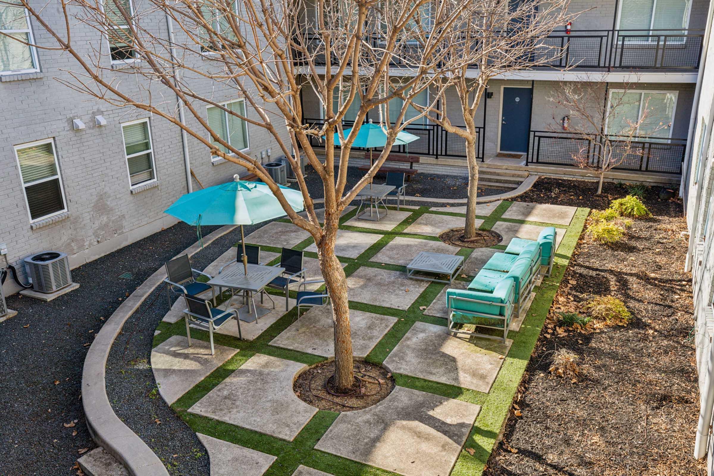A view of a small outdoor courtyard featuring bare trees, turquoise umbrellas over tables, and seating areas surrounded by gravel and grass patches. The courtyard is framed by two buildings with balconies, creating a quiet, inviting space for relaxation.
