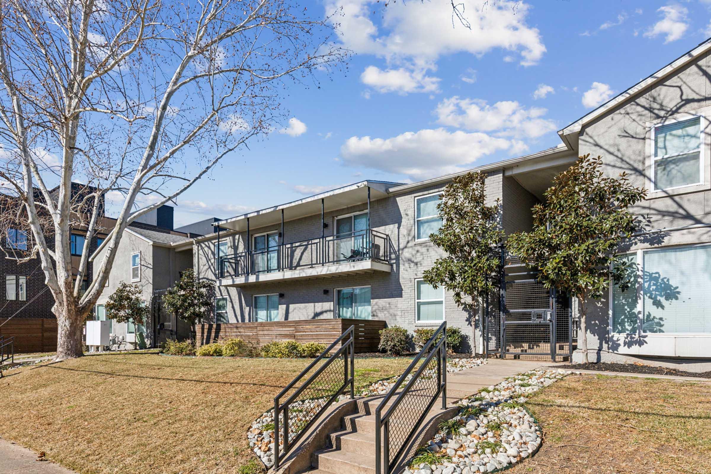 A row of modern multi-story apartment buildings with balconies, surrounded by landscaped grass and trees. A pathway leads up to the entrance, with decorative rocks lining the steps. The sky is partly cloudy, indicating a bright day.