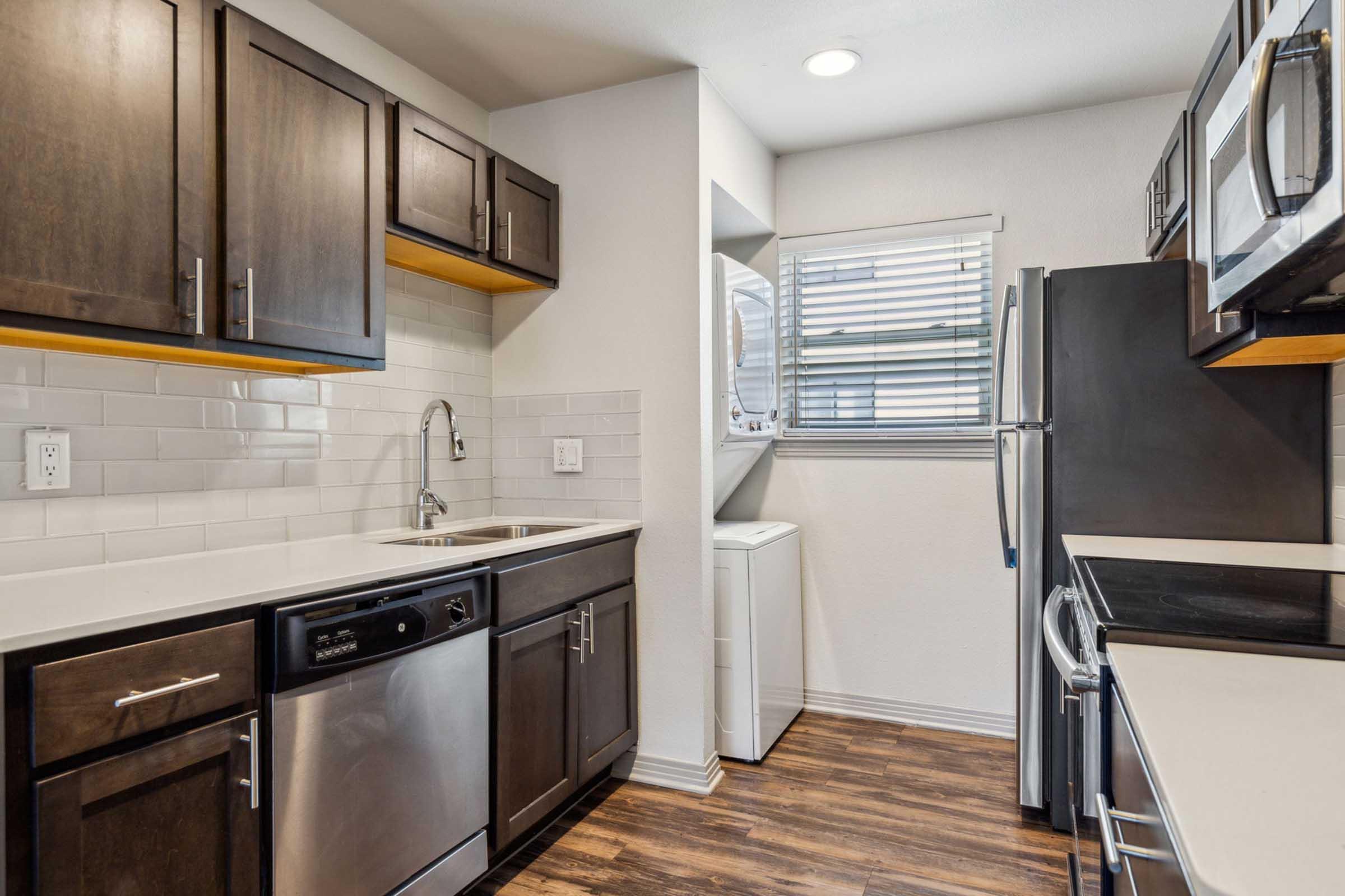 Modern kitchen with dark wood cabinets, stainless steel appliances, and white countertops. Includes a sink, dishwasher, and microwave, along with a laundry area featuring a washing machine. Bright natural light comes from a window, complemented by sleek, laminate flooring.